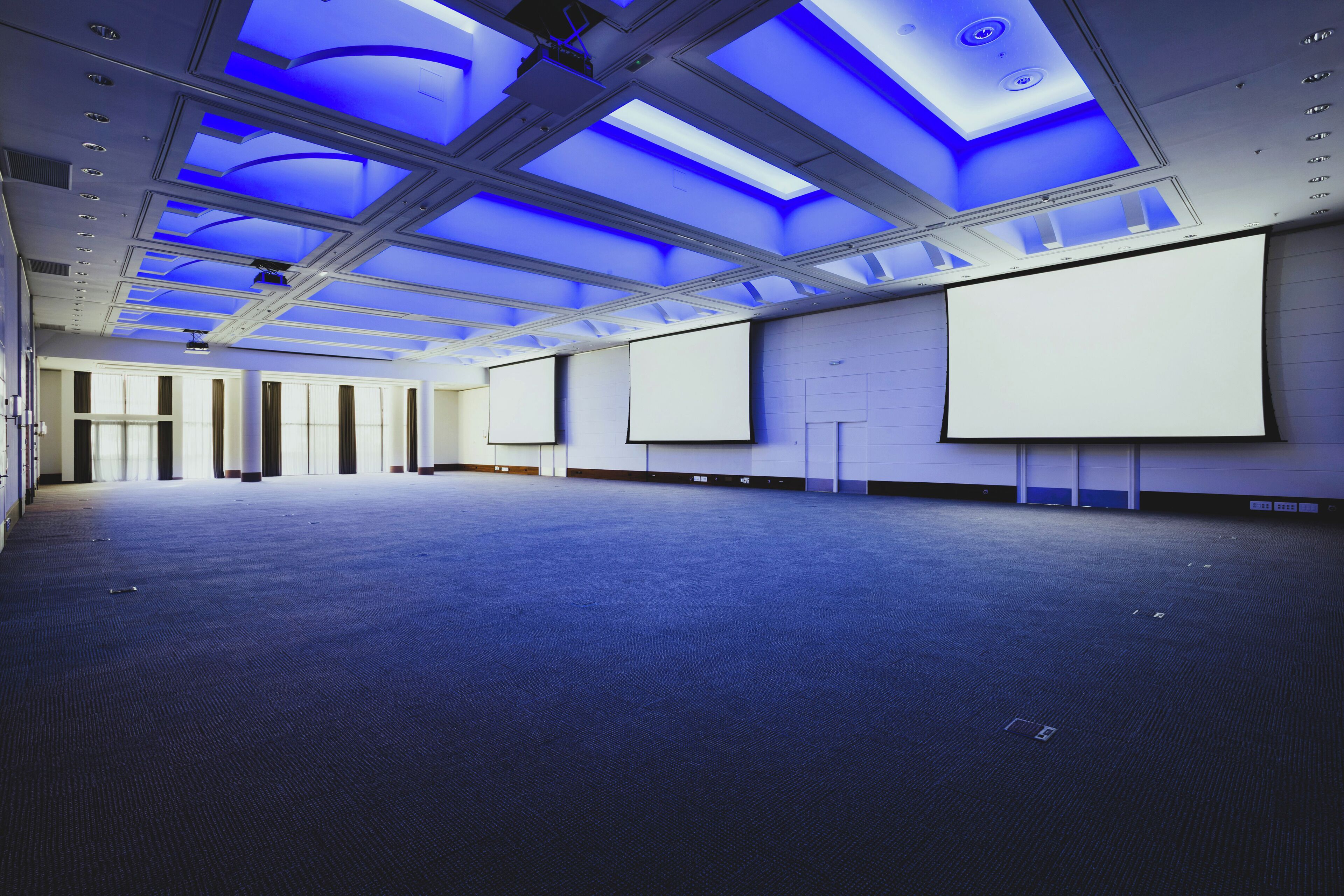 A large, empty conference room with a high ceiling, skylights, and multiple projection screens.