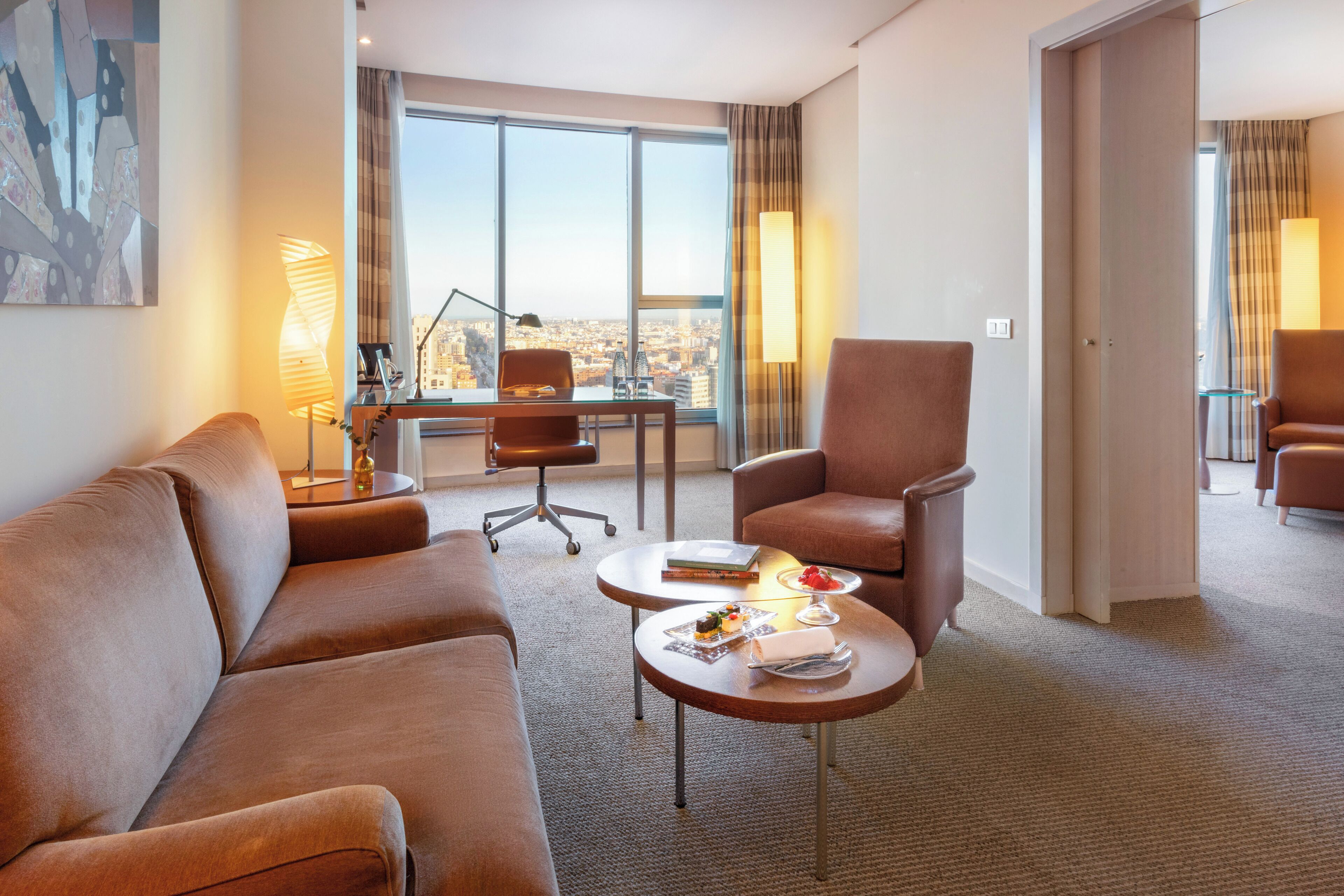 Una habitación de hotel moderna con un ventanal grande, un sofá, una silla, una mesita con un libro y un vaso, y una lámpara.