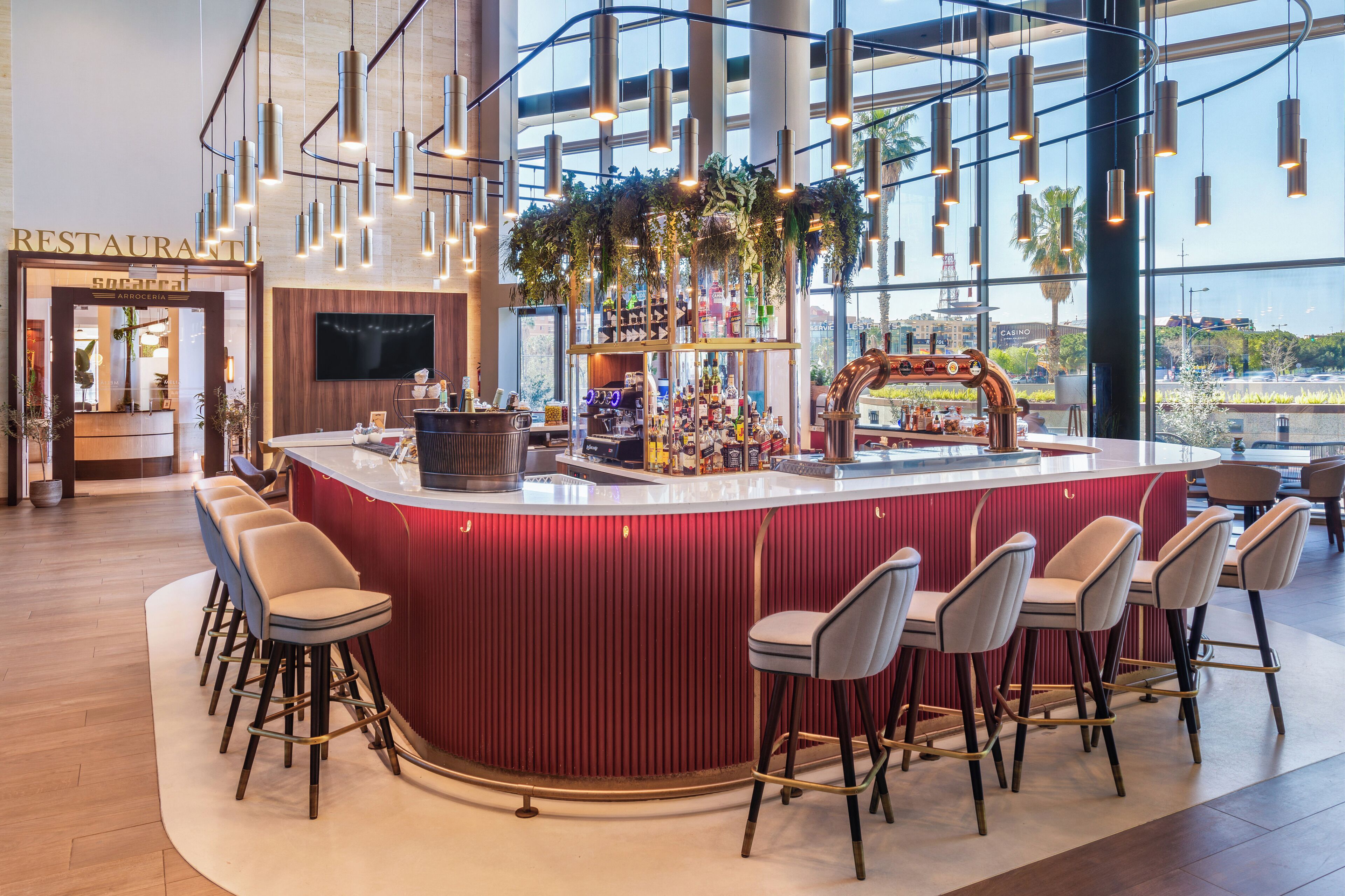 A modern bar area with a red bar counter, multiple bar stools, and a large window offering a view of the city.