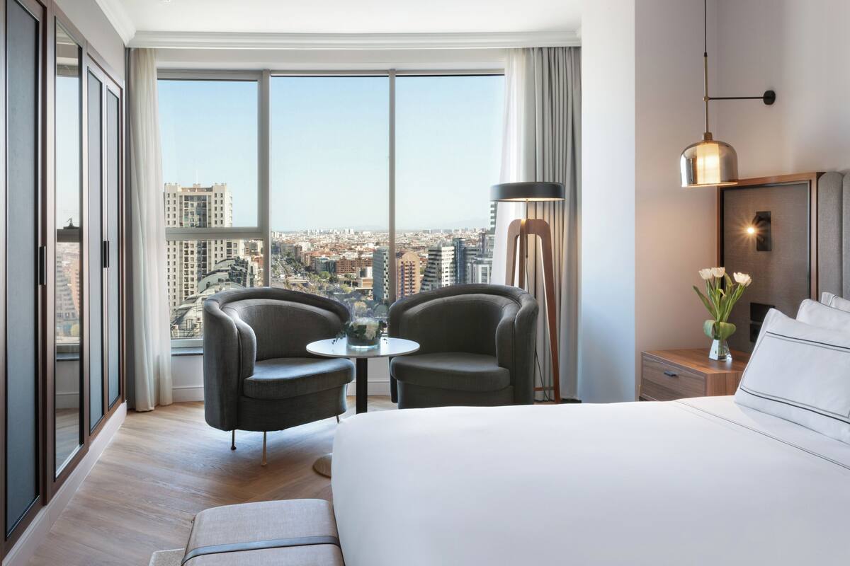 Habitación de hotel con un ventanal grande, dos sillones, una mesita, una cama, una mesita de noche y una lámpara.