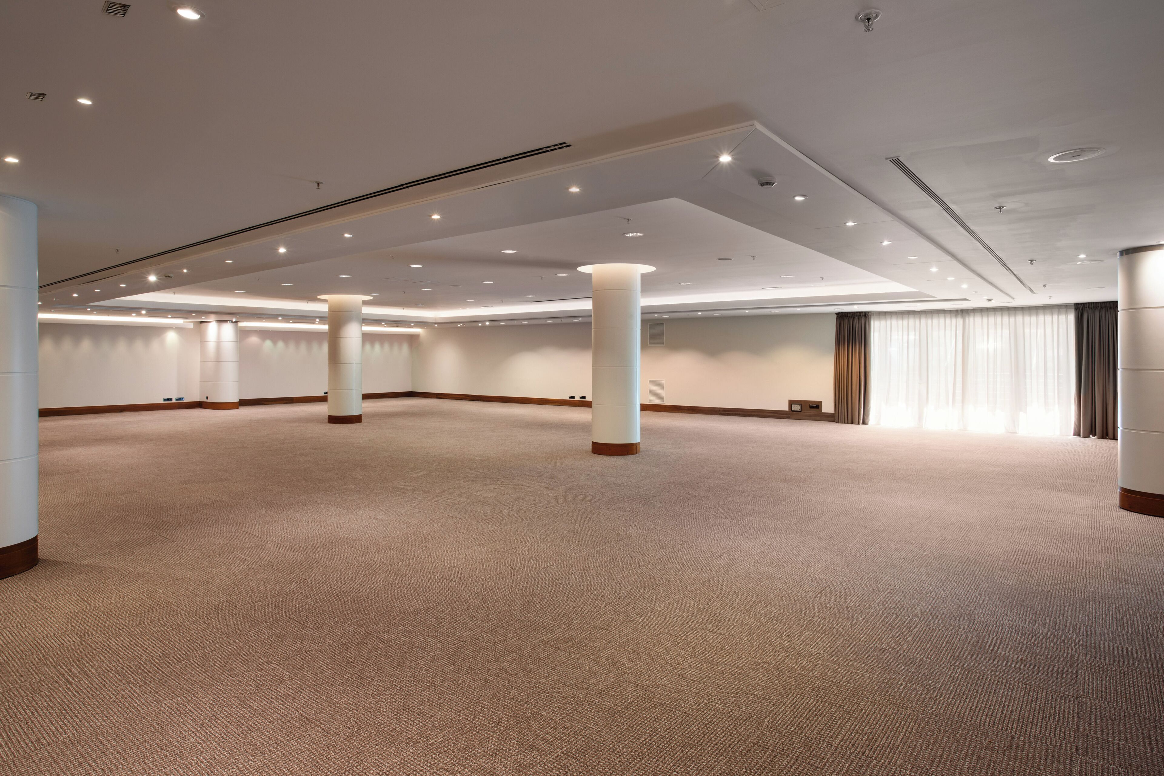 A spacious, well-lit conference room with a high ceiling, carpeted floor, and large windows with curtains.