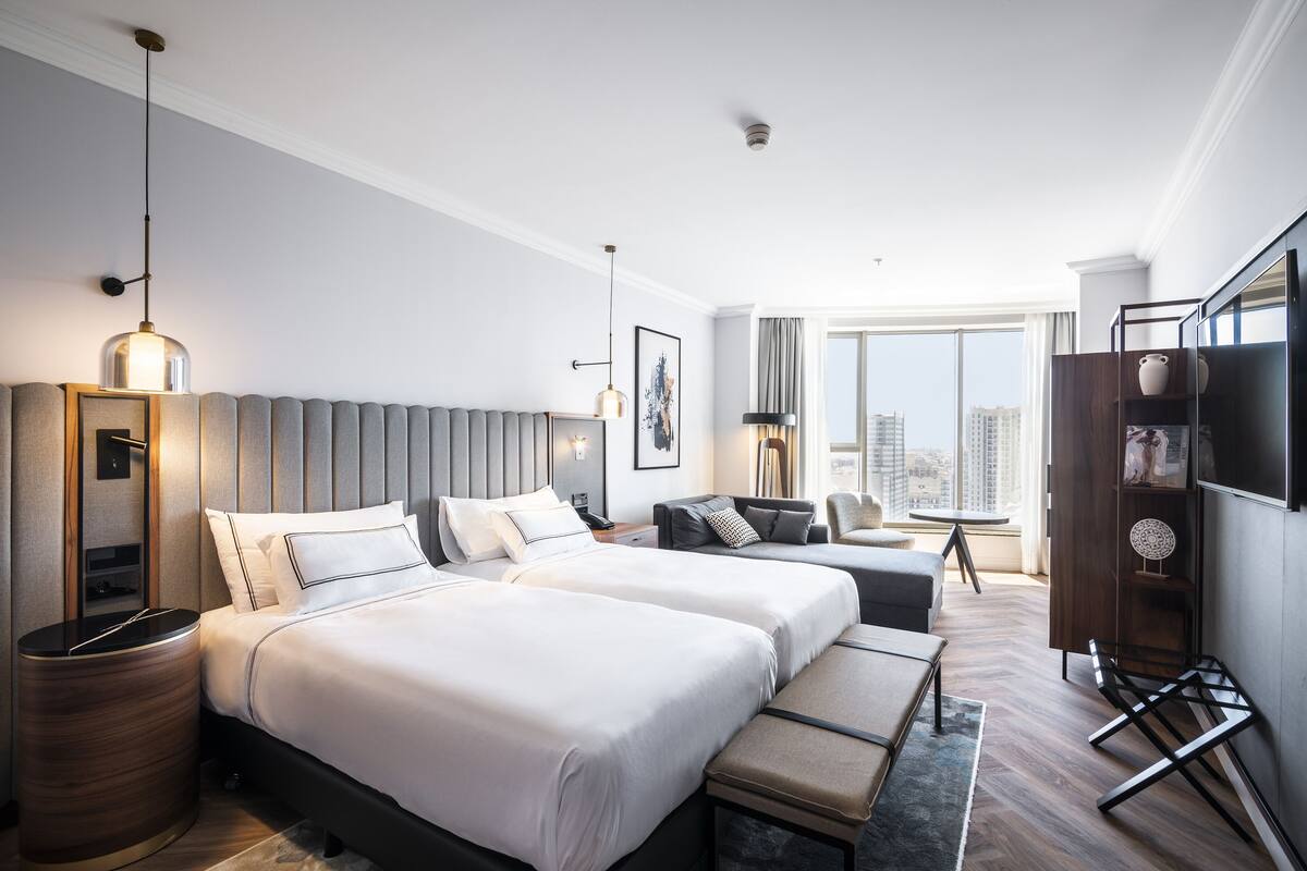 A hotel room with two beds, a sofa, a TV, and a large window with a city view.