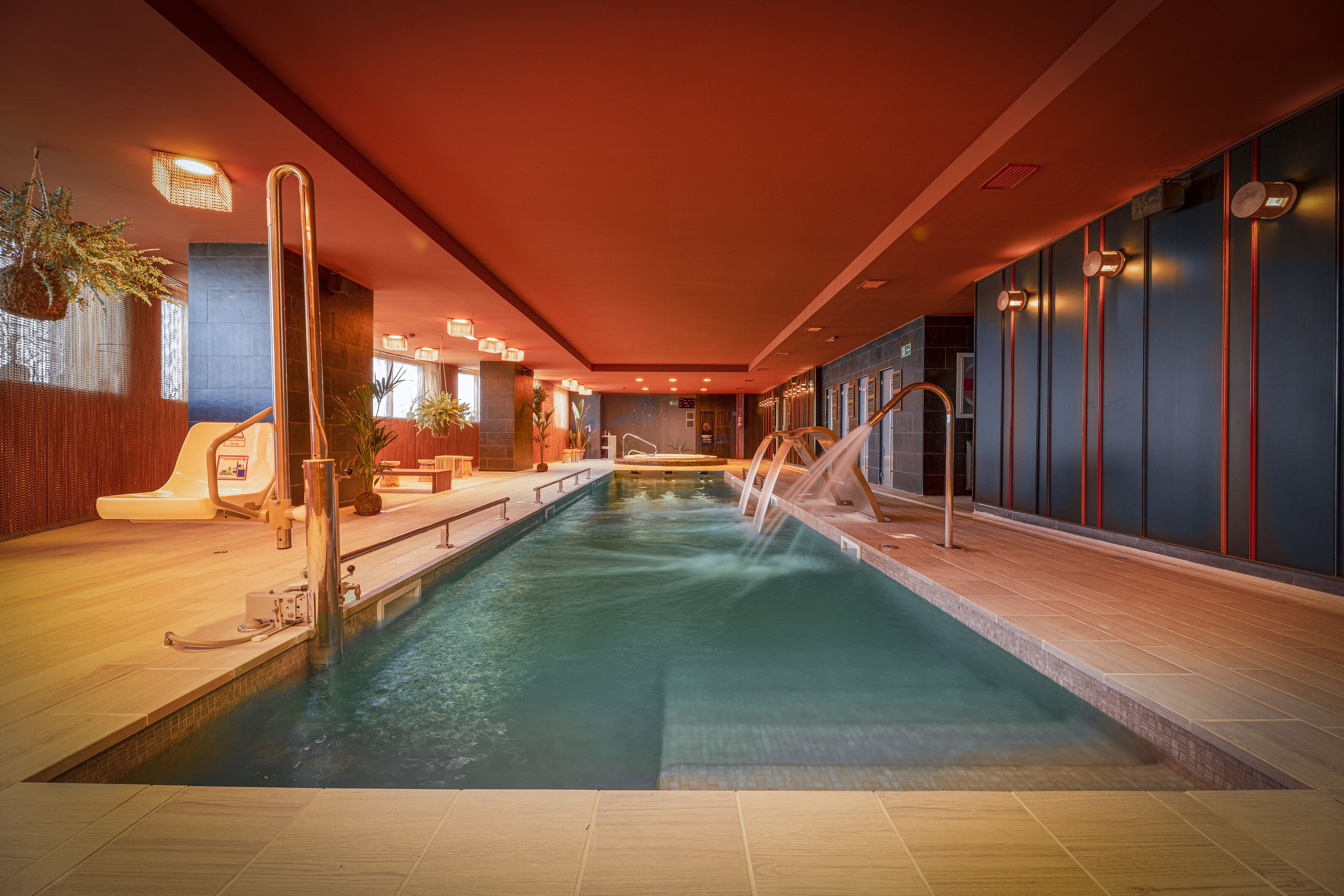 An indoor swimming pool with a modern design, featuring a long, narrow pool, a central water feature, and a seating area with a sun lounger.