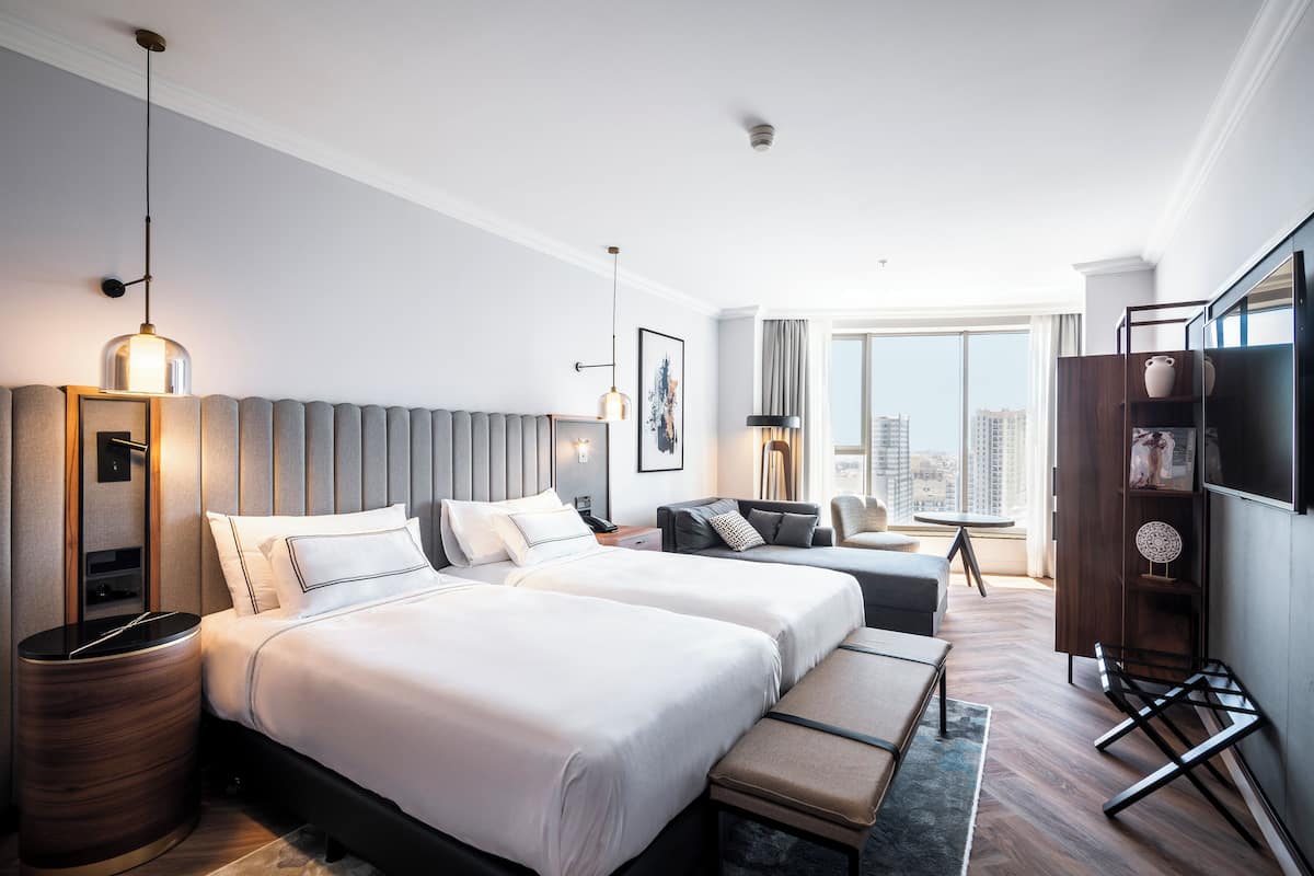 Quarto de hotel com duas camas, televisão, secretária e uma janela grande com vista para a cidade.