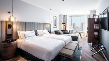 Habitación de hotel con dos camas, televisión, escritorio y ventana grande con vistas a la ciudad.