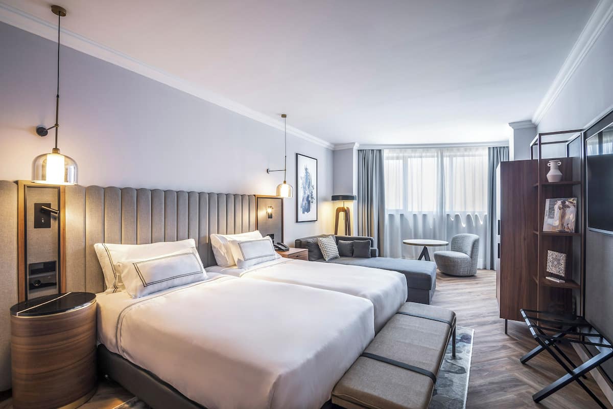 Quarto de hotel com duas camas, um sofá, uma mesa pequena, uma cadeira, uma televisão e uma estante de livros.