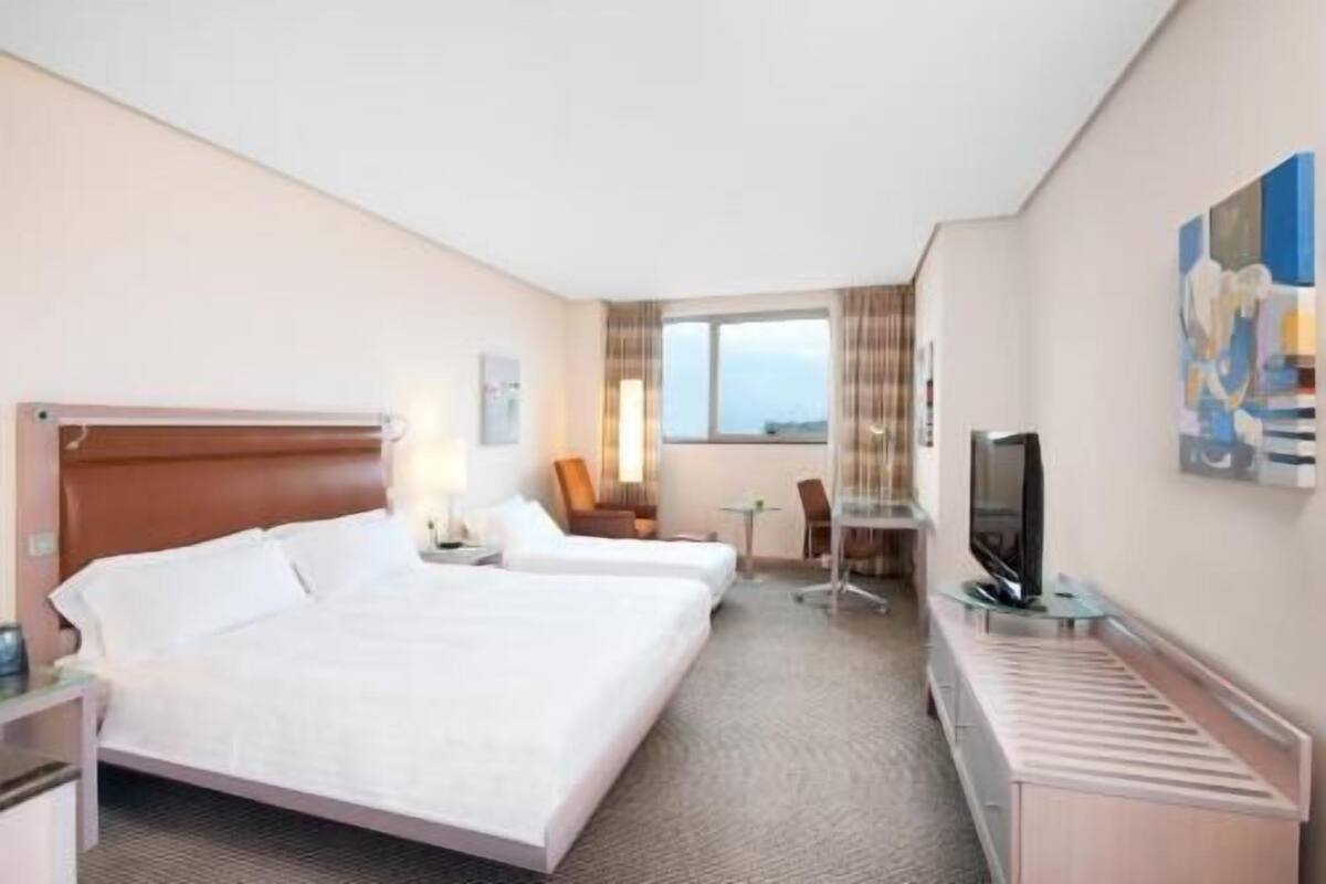 Quarto de hotel com duas camas, uma escrivaninha, uma cadeira, uma televisão e uma janela.