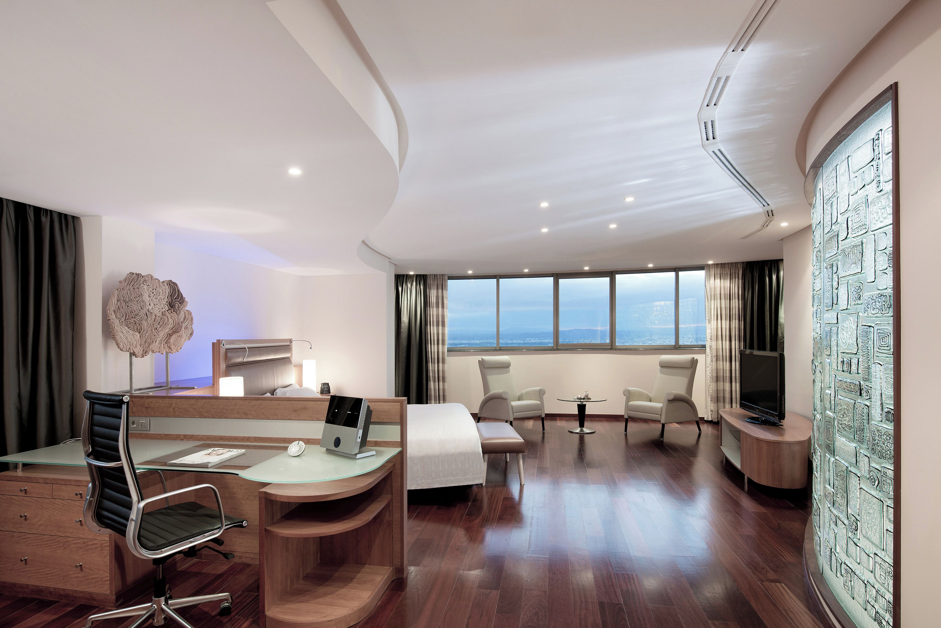 Ein Hotelzimmer mit Schreibtisch, Bett, Fernseher und Blick auf die Stadt.