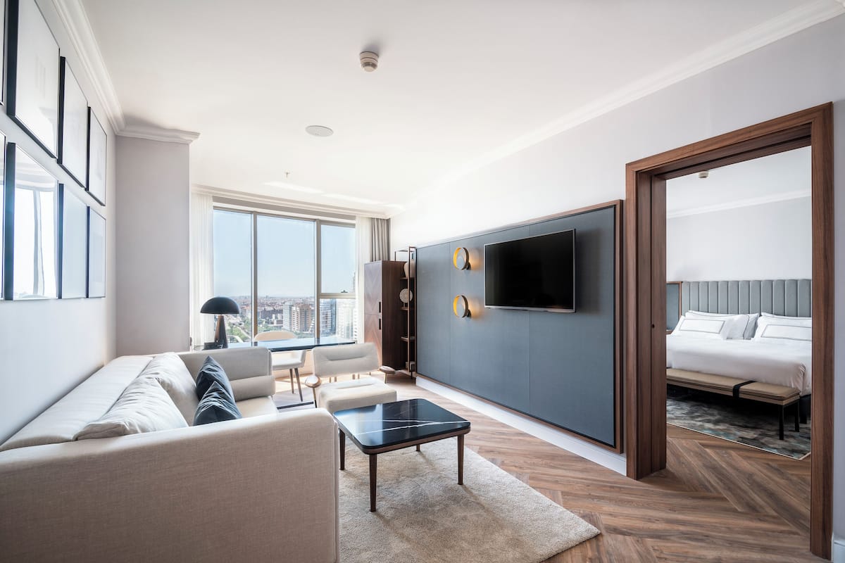 Quarto de hotel moderno com uma cama grande, um sofá, uma mesa de centro, uma TV fixada na parede e vista para a cidade.
