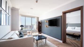 Quarto de hotel moderno com uma cama grande, um sofá, uma mesa de centro, uma TV fixada na parede e vista para a cidade.