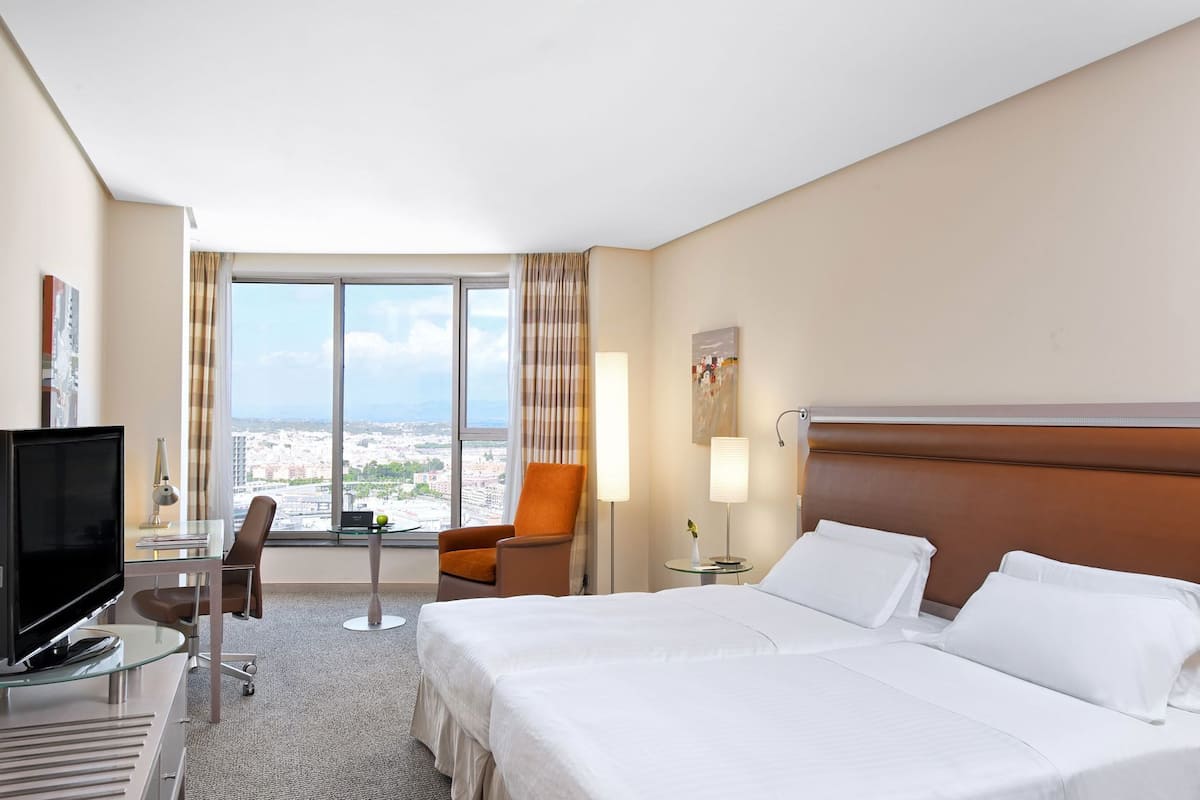 Quarto de hotel com cama, escrivaninha com televisão, cadeira, abajur e vista da cidade.