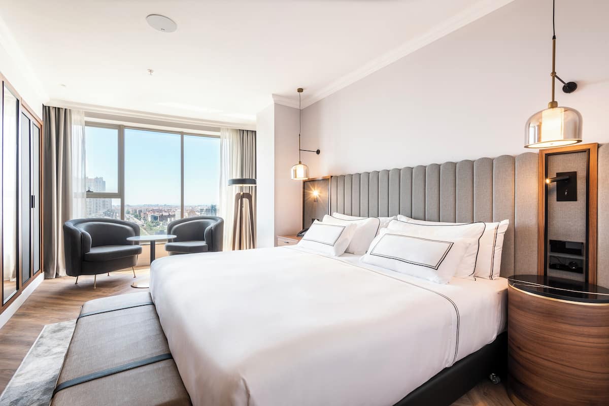 Quarto de hotel moderno com uma cama grande, área de estar com duas cadeiras, vista da cidade e uma janela ampla.