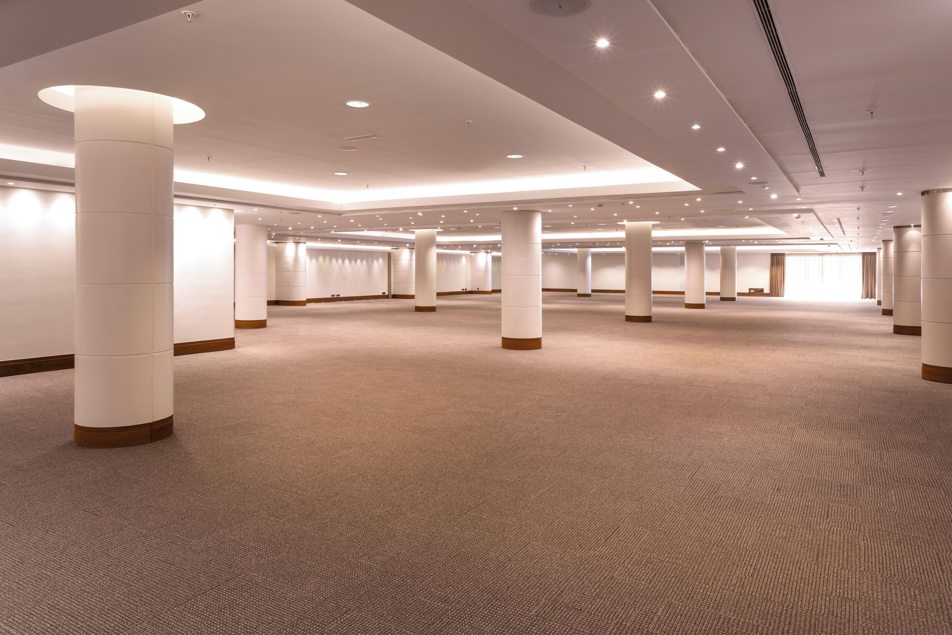 Amplia habitación de hotel bien iluminada, con piso alfombrado, grandes pilares y un diseño moderno en el techo.
