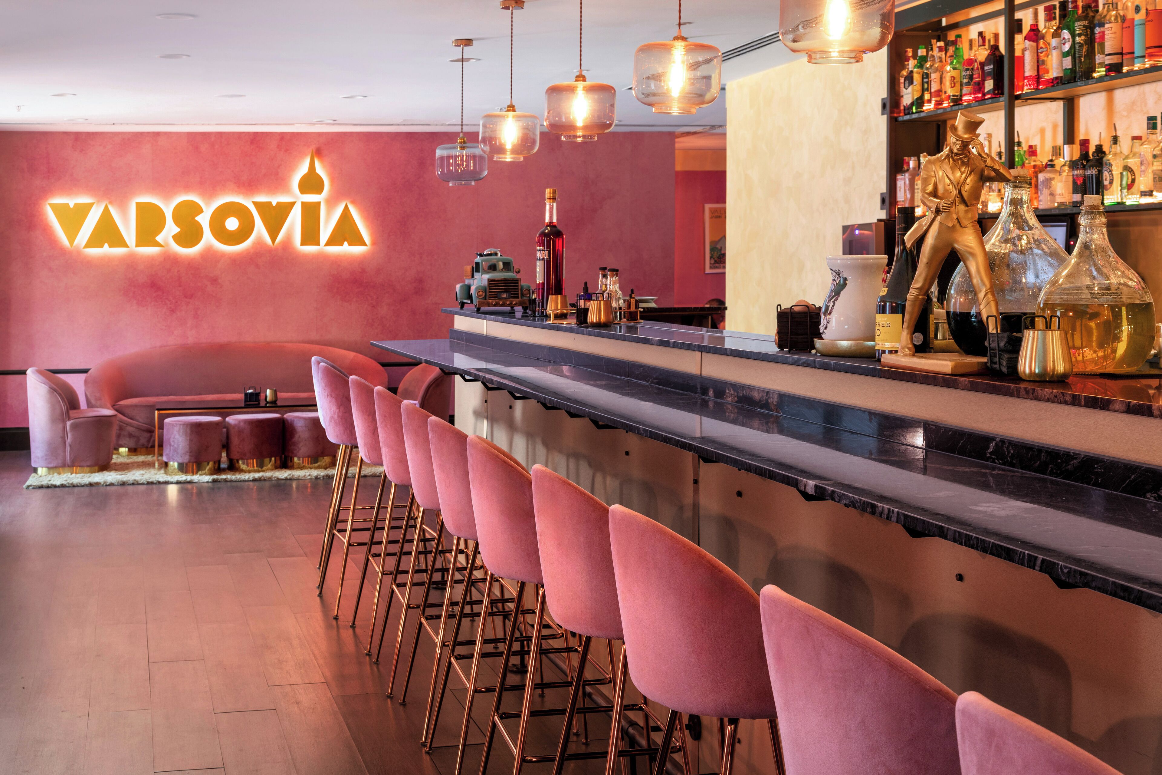 Una zona de bar con un letrero de neón que dice "VARSOVIA", una barra larga con taburetes rosados y una estatua en la estantería.