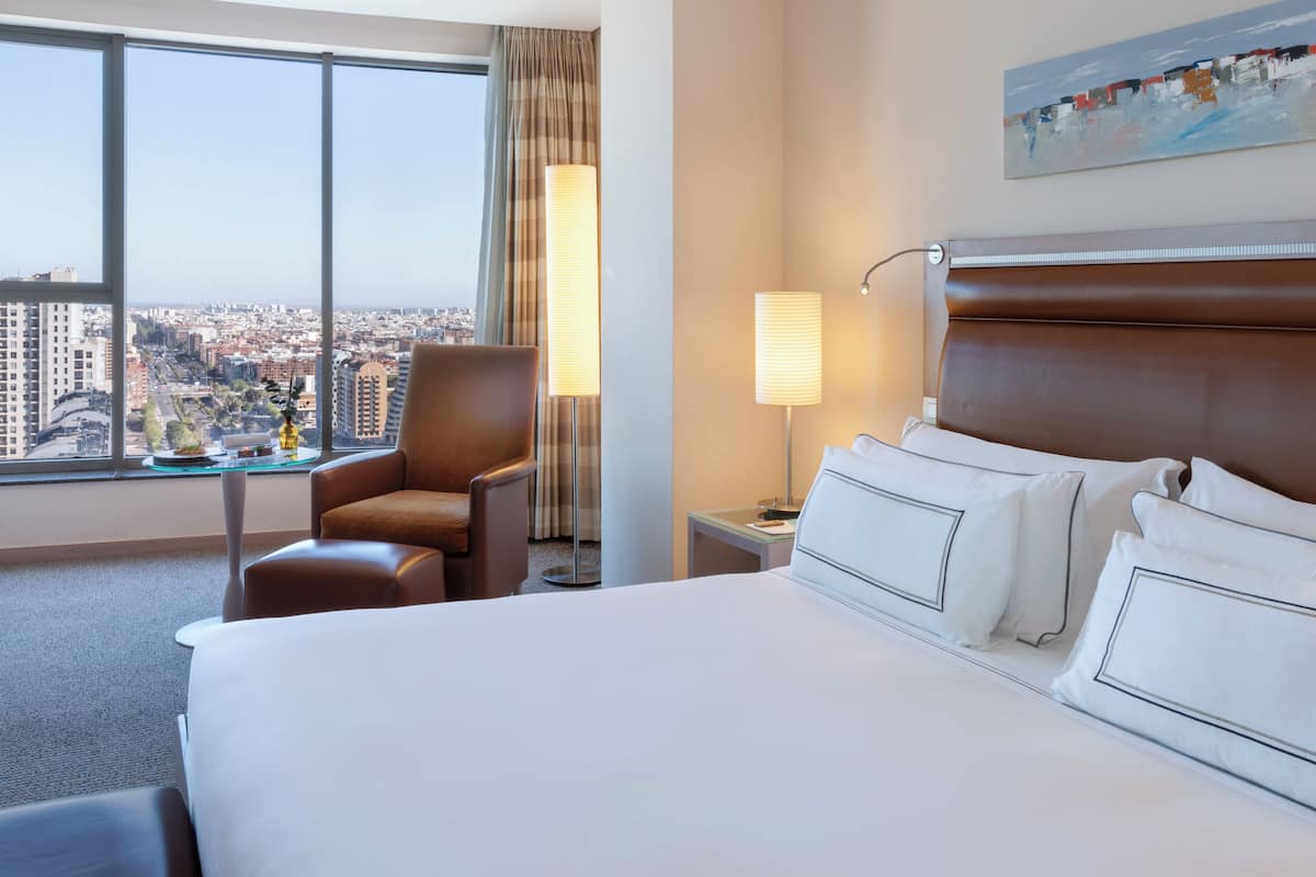 Quarto de hotel com uma cama grande, uma cadeira, uma mesa de cabeceira, um abajur e vista para a cidade através de janelas amplas.