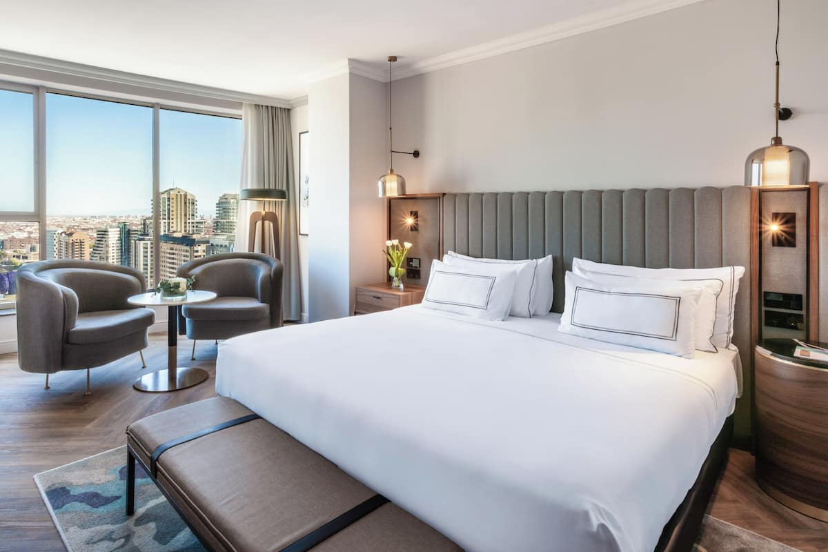 Quarto de hotel com uma cama grande, duas poltronas, uma mesinha e vista para a cidade.