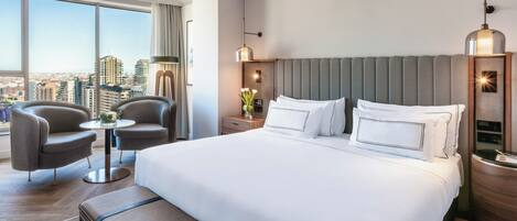 A hotel room with a large bed, two armchairs, a small table, and a view of the city.
