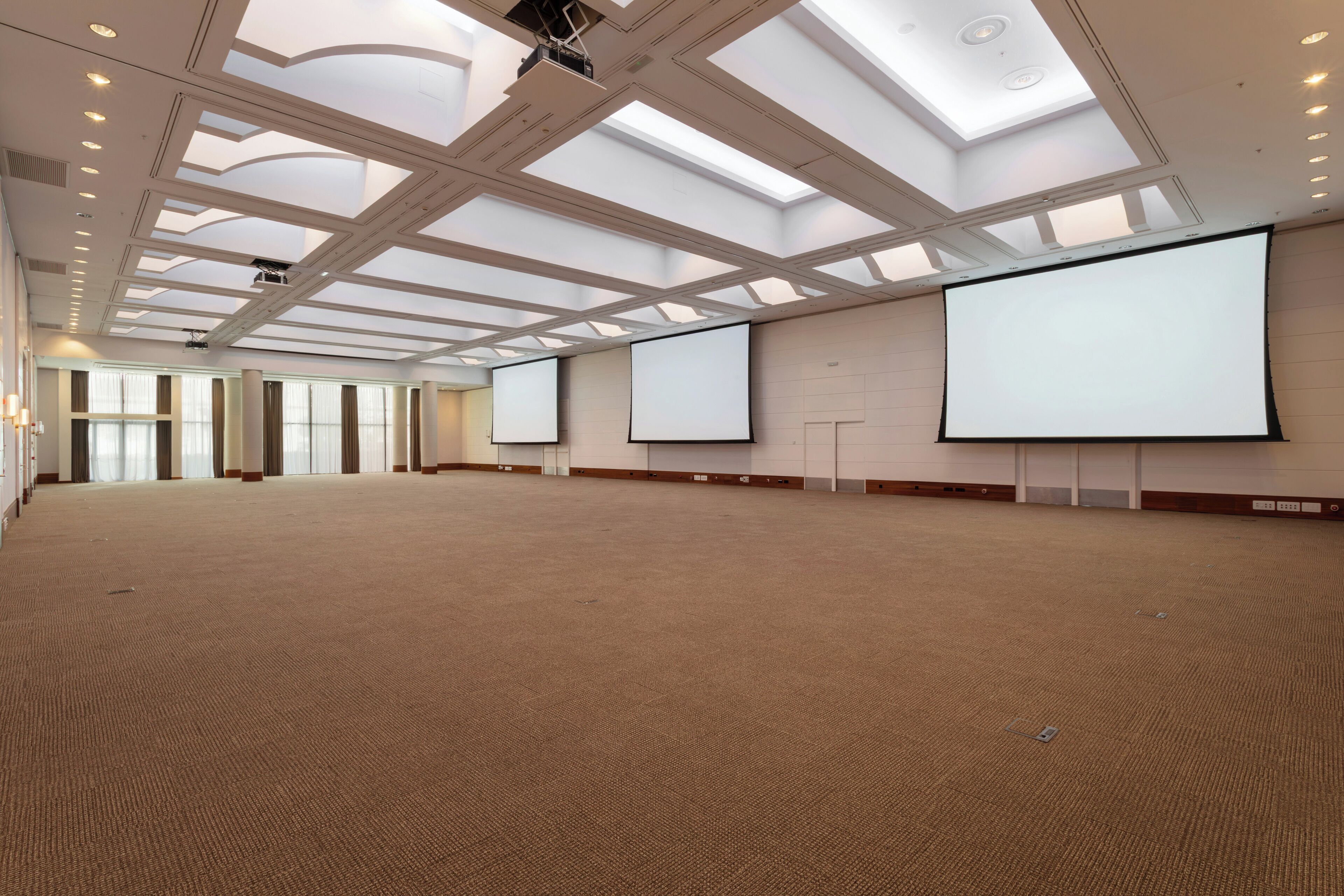 Un amplio salón de conferencias bien iluminado, con techos altos, tragaluces y varias pantallas de proyección.