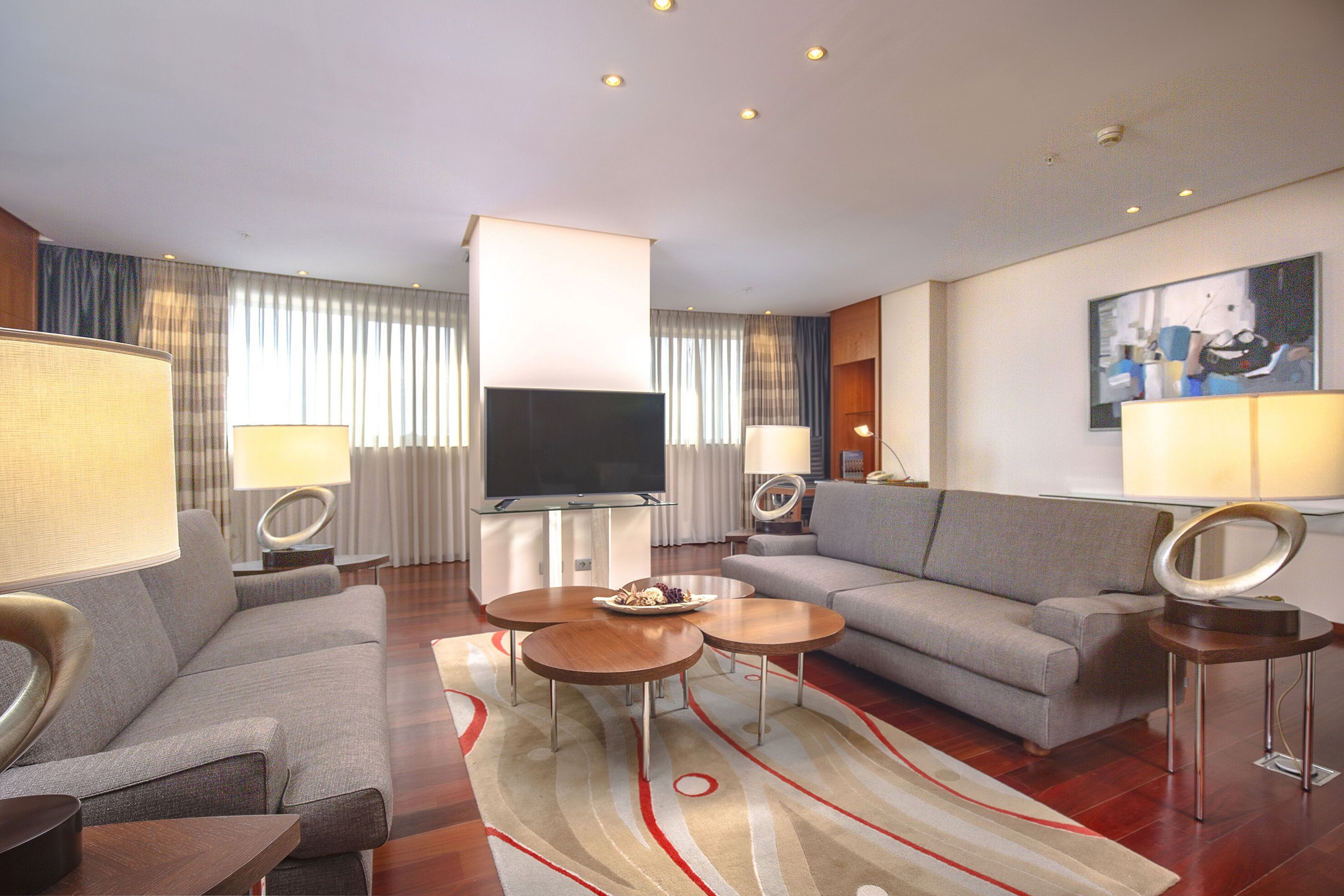 A modern hotel room with a grey sofa, a flat-screen TV, two side tables with lamps, and a large painting on the wall.