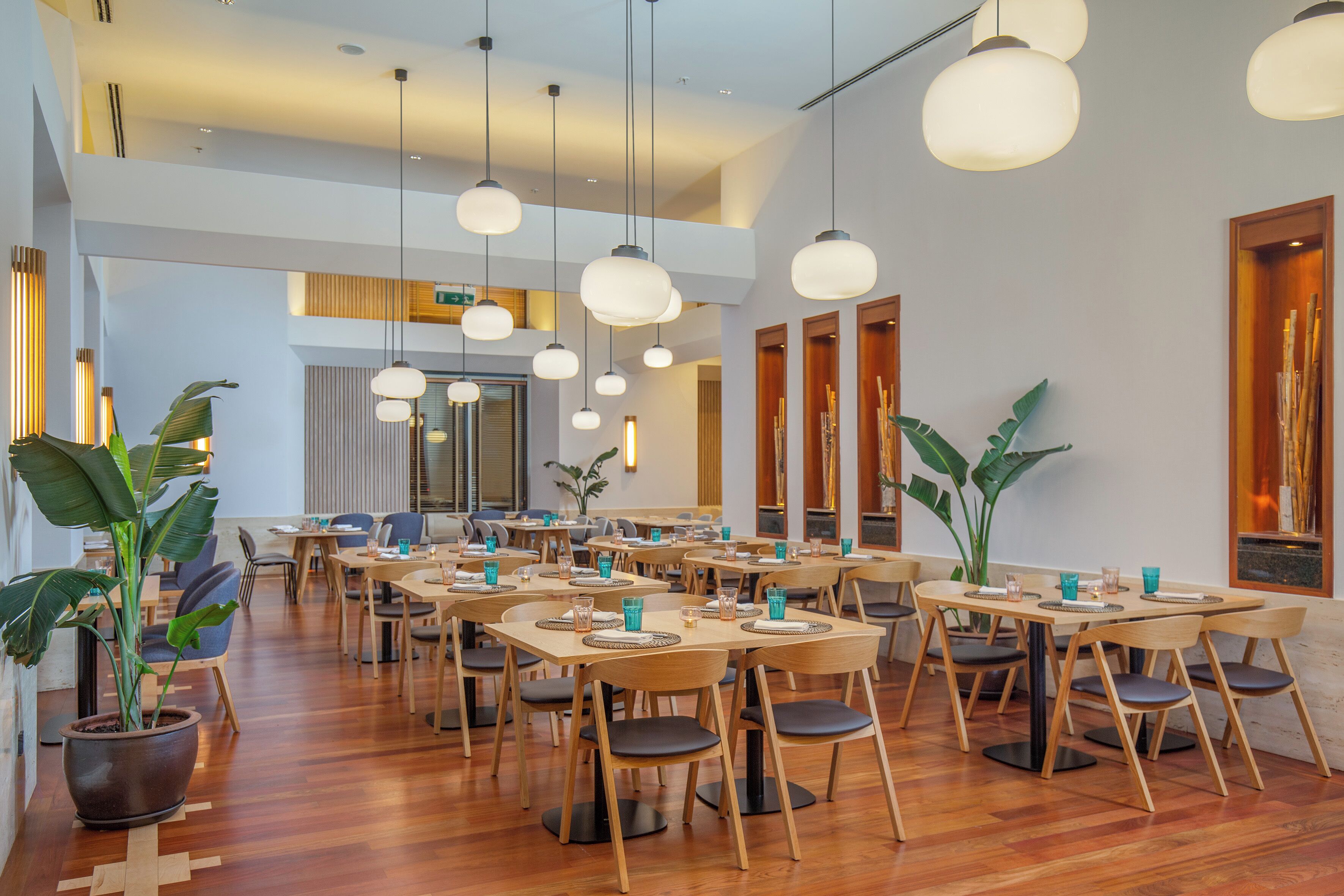 Un comedor amplio con varias mesas y sillas, piso de madera y lámparas colgantes.