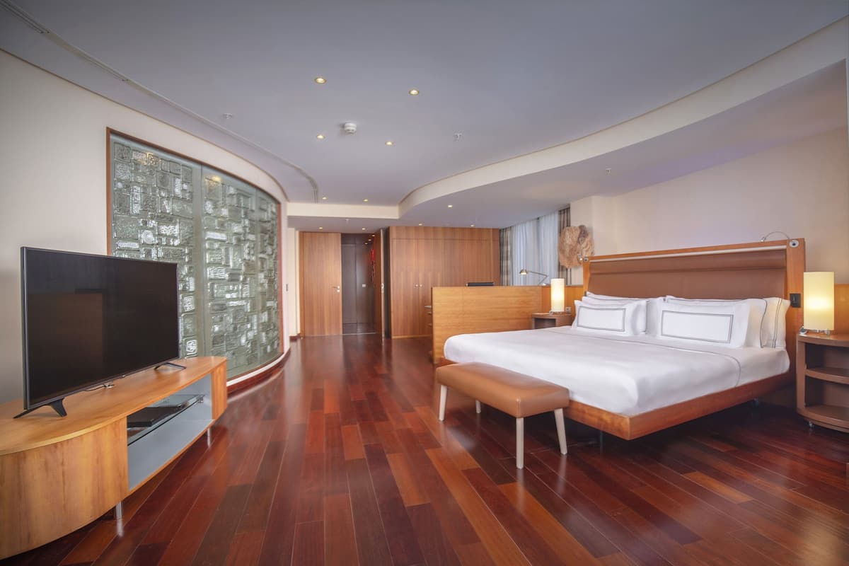 Quarto de hotel moderno com uma cama grande, televisão de tela plana, piso de madeira e parede de vidro com vista para a cidade.
