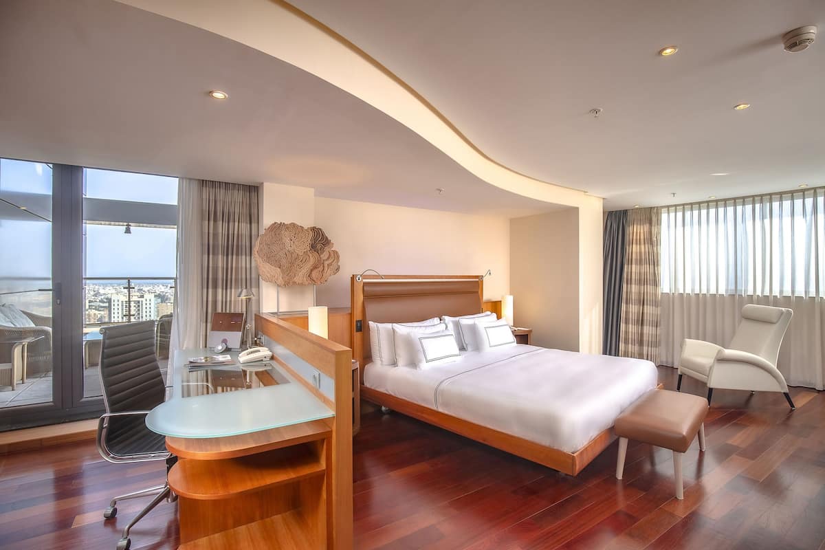 Quarto de hotel moderno com uma cama grande, uma mesa e uma cadeira. Há vista da cidade através de amplas janelas.