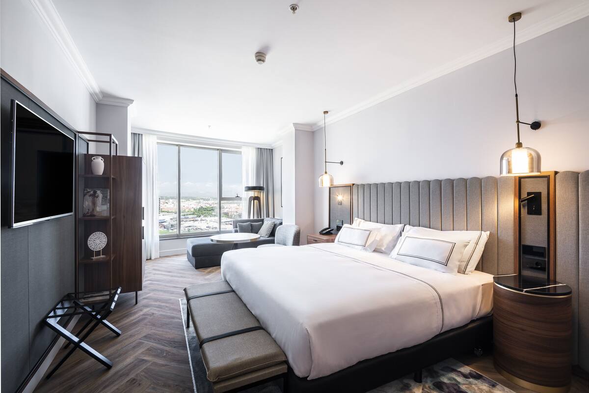 Quarto de hotel moderno com uma cama grande, área de estar com sofá e poltronas, televisão e uma janela ampla com vista para a cidade.