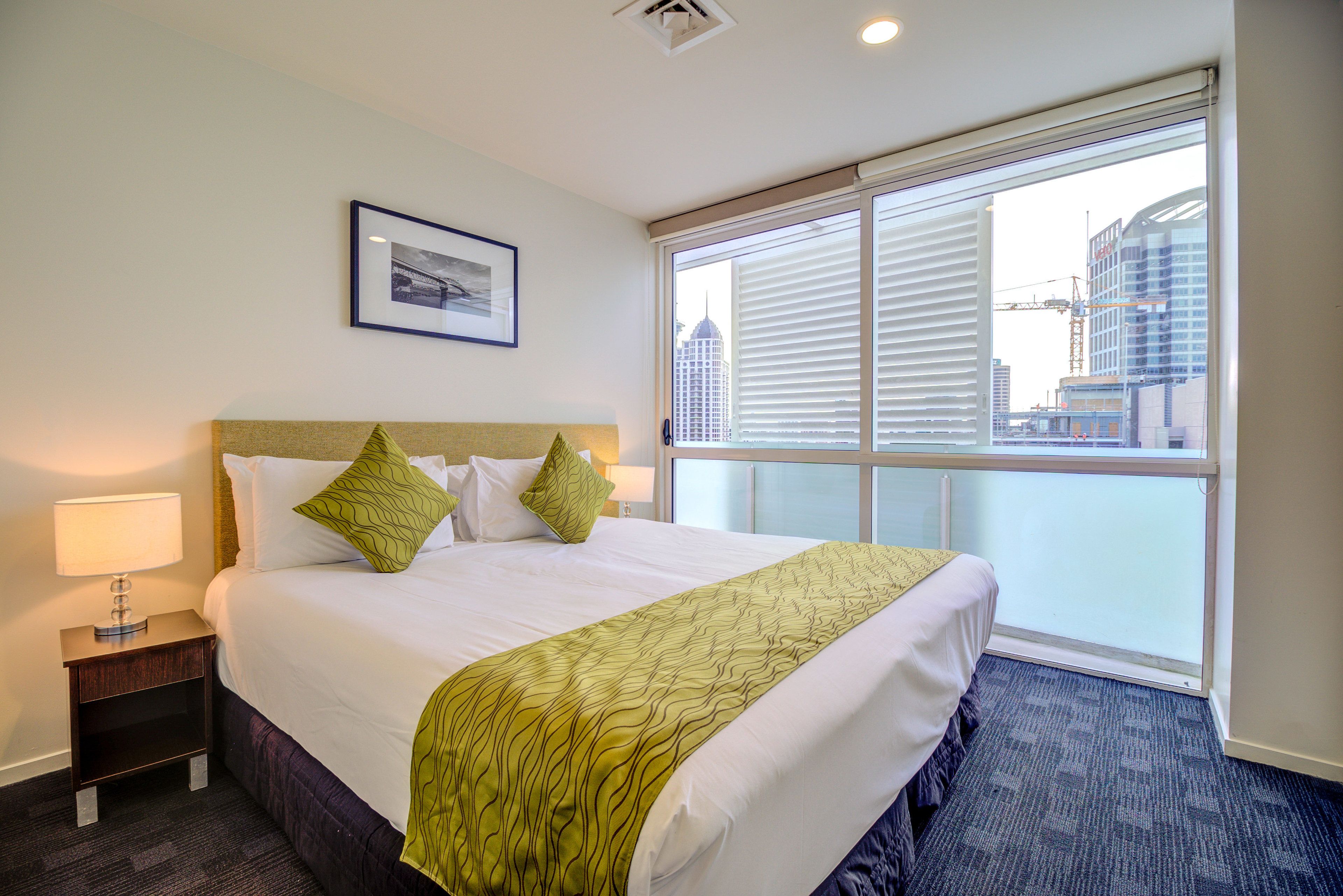 A hotel room with a large bed, a nightstand with a lamp, a framed picture on the wall, and a view of the city through a large window.