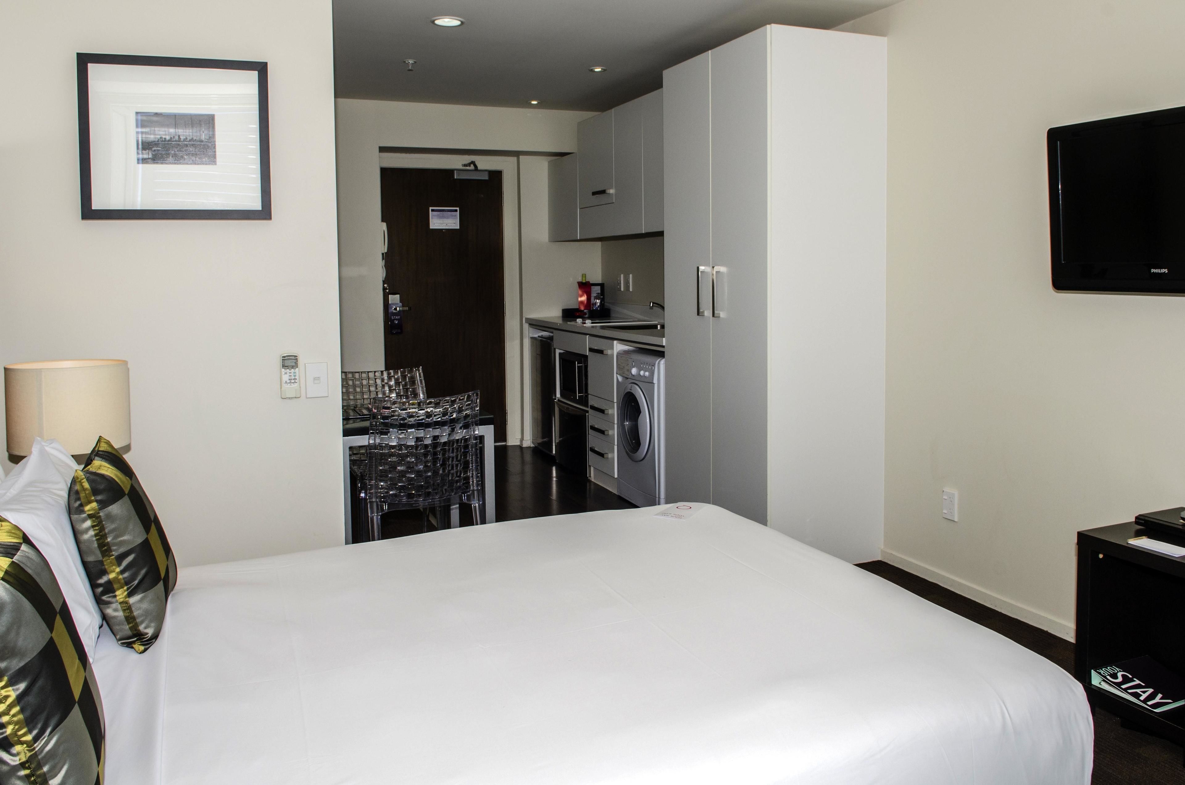 A hotel room with a bed, a TV, a desk, a lamp, and a kitchenette with a washing machine.