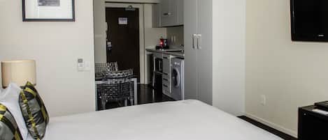 A hotel room with a bed, a TV, a desk, a lamp, and a kitchenette with a washing machine.