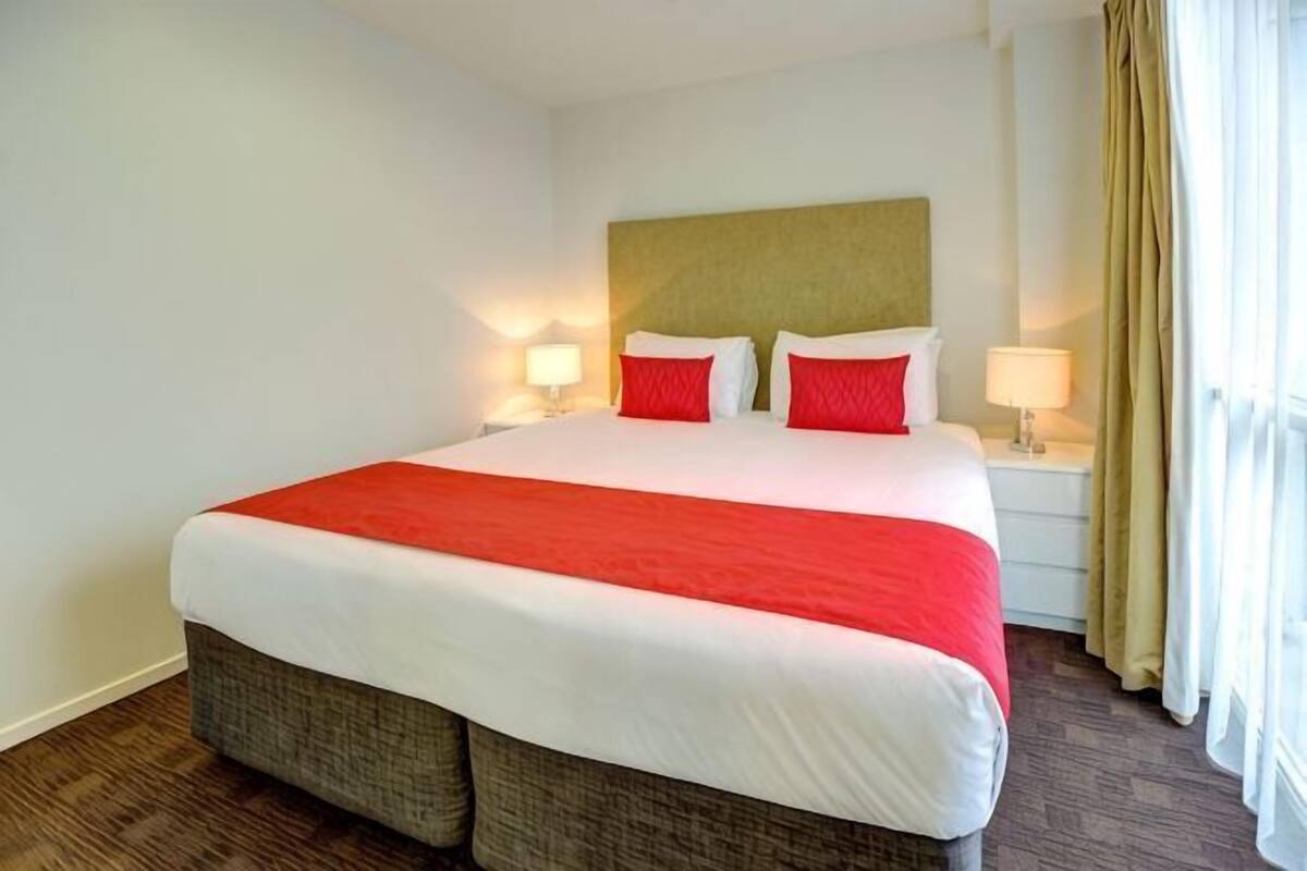 A hotel room with a large bed, two bedside tables with lamps, a headboard, and a window with curtains.