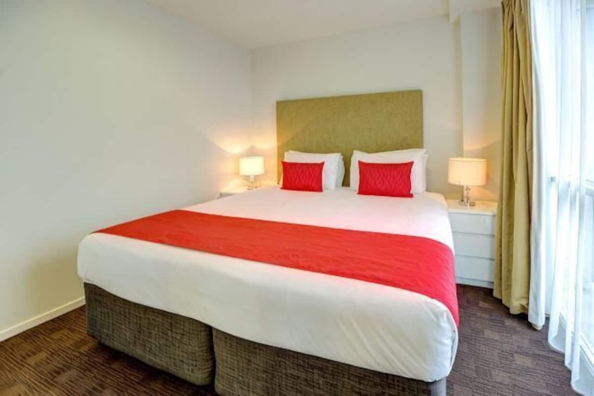 Una habitación de hotel con una cama grande, dos mesitas de noche con lámparas, un cabecero y una ventana con cortinas.