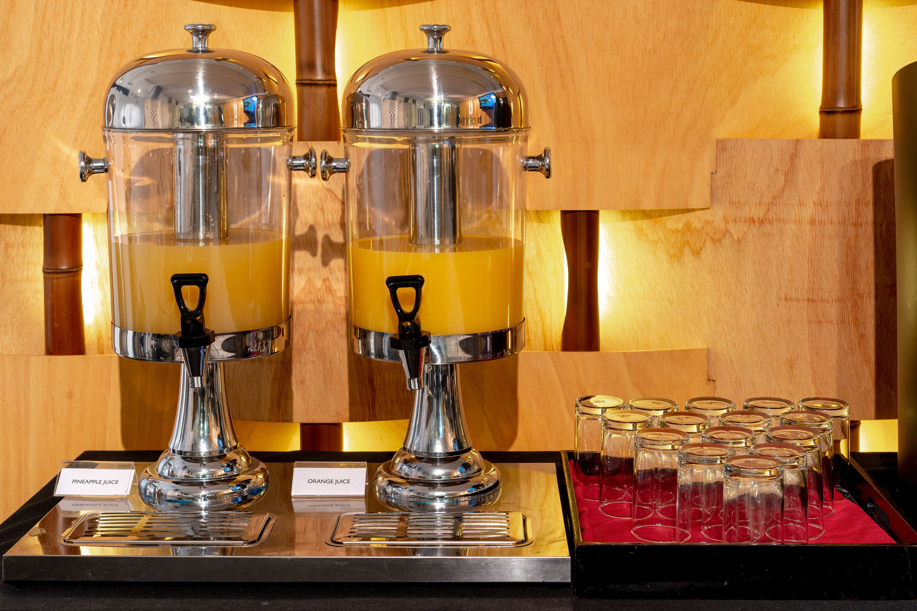Two stainless steel beverage dispensers with transparent containers, labeled for pineapple and orange juice, and a tray of empty glasses.