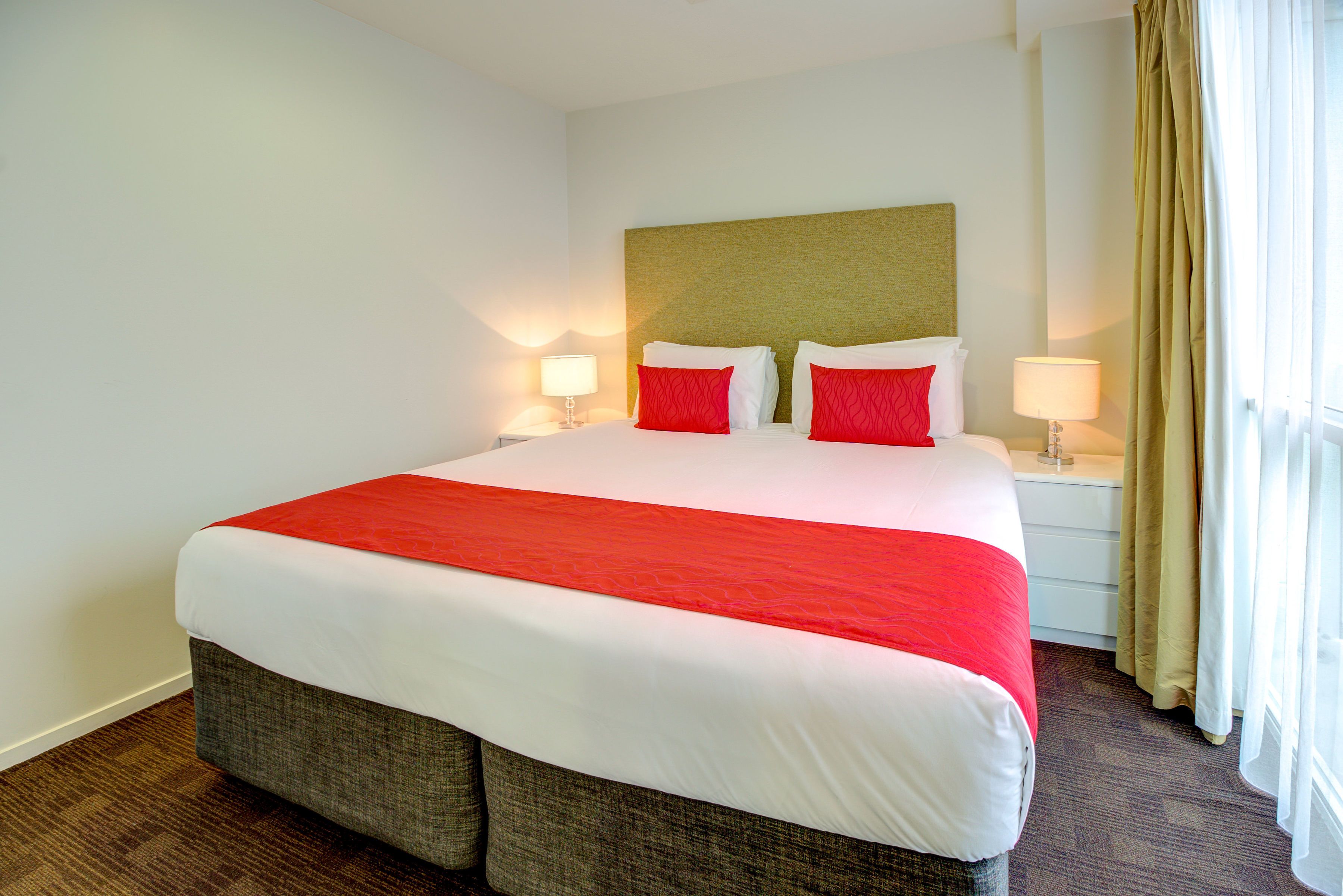 A hotel room with a large bed, two bedside tables with lamps, a headboard, and a window with curtains.