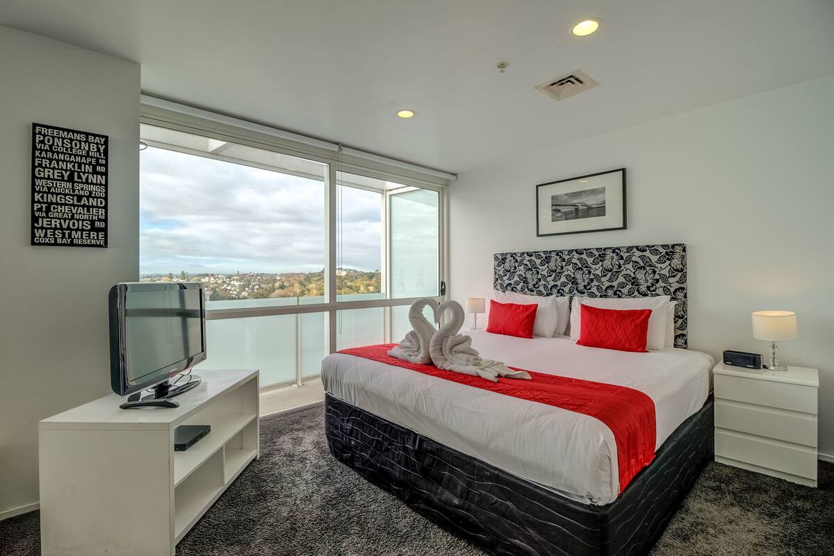 A hotel room with a large bed, a TV on a white stand, a bedside table with a lamp, and a view of the city through large windows.
