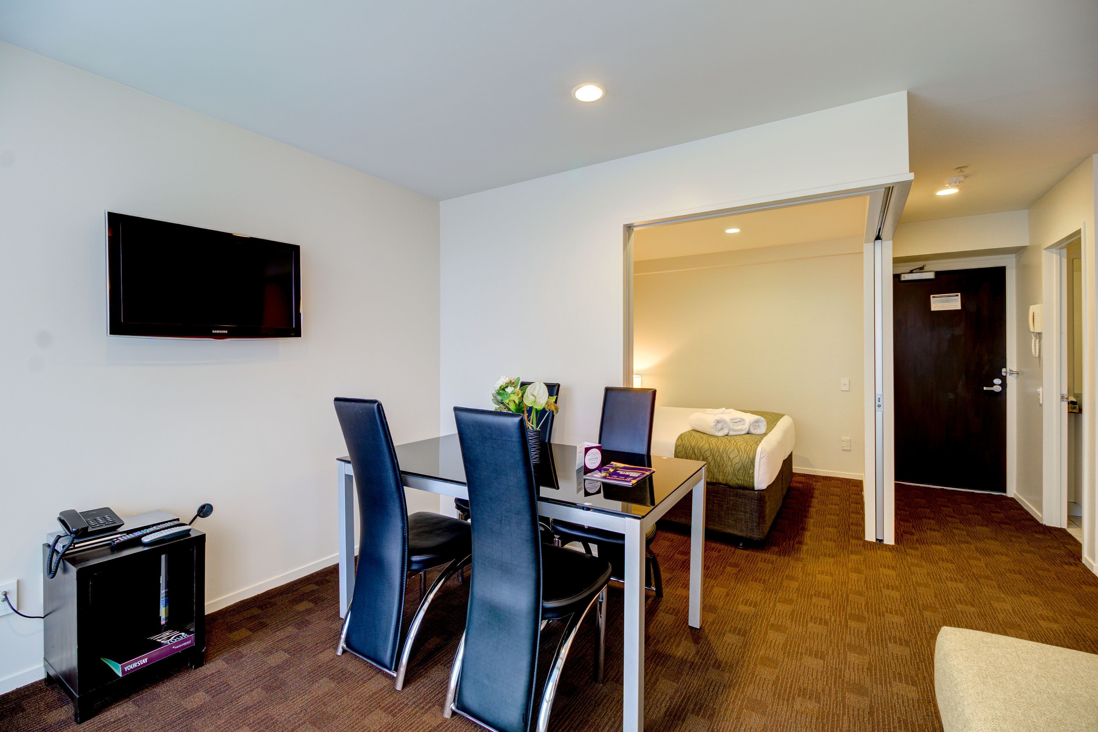 A hotel room with a bed, a desk with a computer, a TV mounted on the wall, and a door leading to another room.