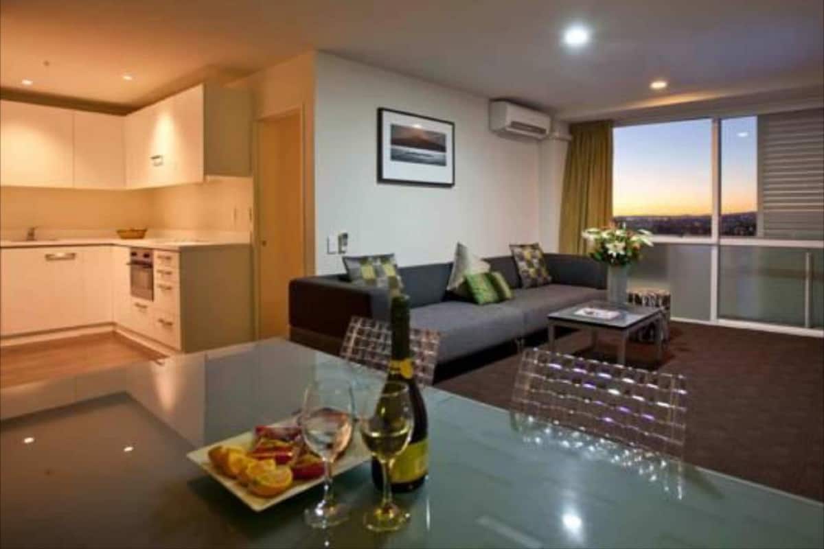 A modern living room with a sofa, a dining table with a plate of food and wine, and a view of the city at sunset.