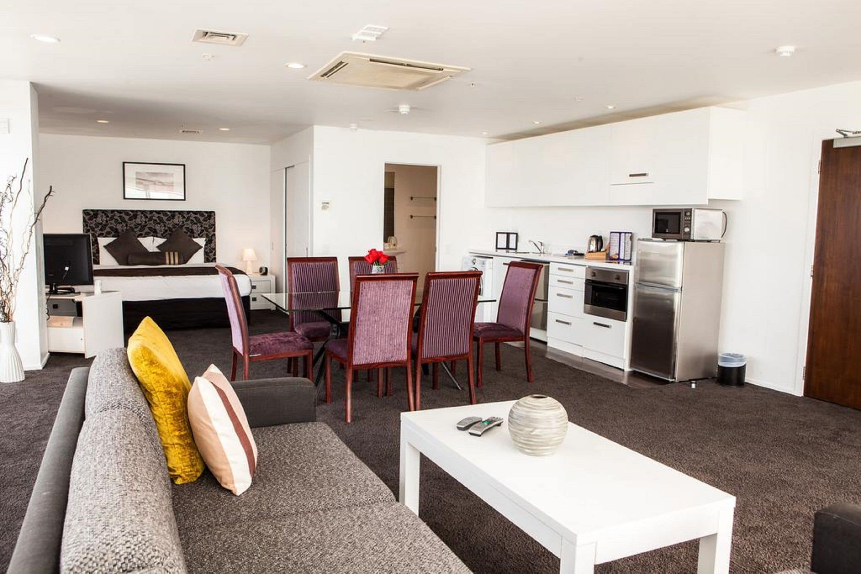 A modern living room with a grey sofa, a white coffee table, and a dining area with a red flower centerpiece. A kitchen with white cabinets and stainless steel appliances is visible in the background.