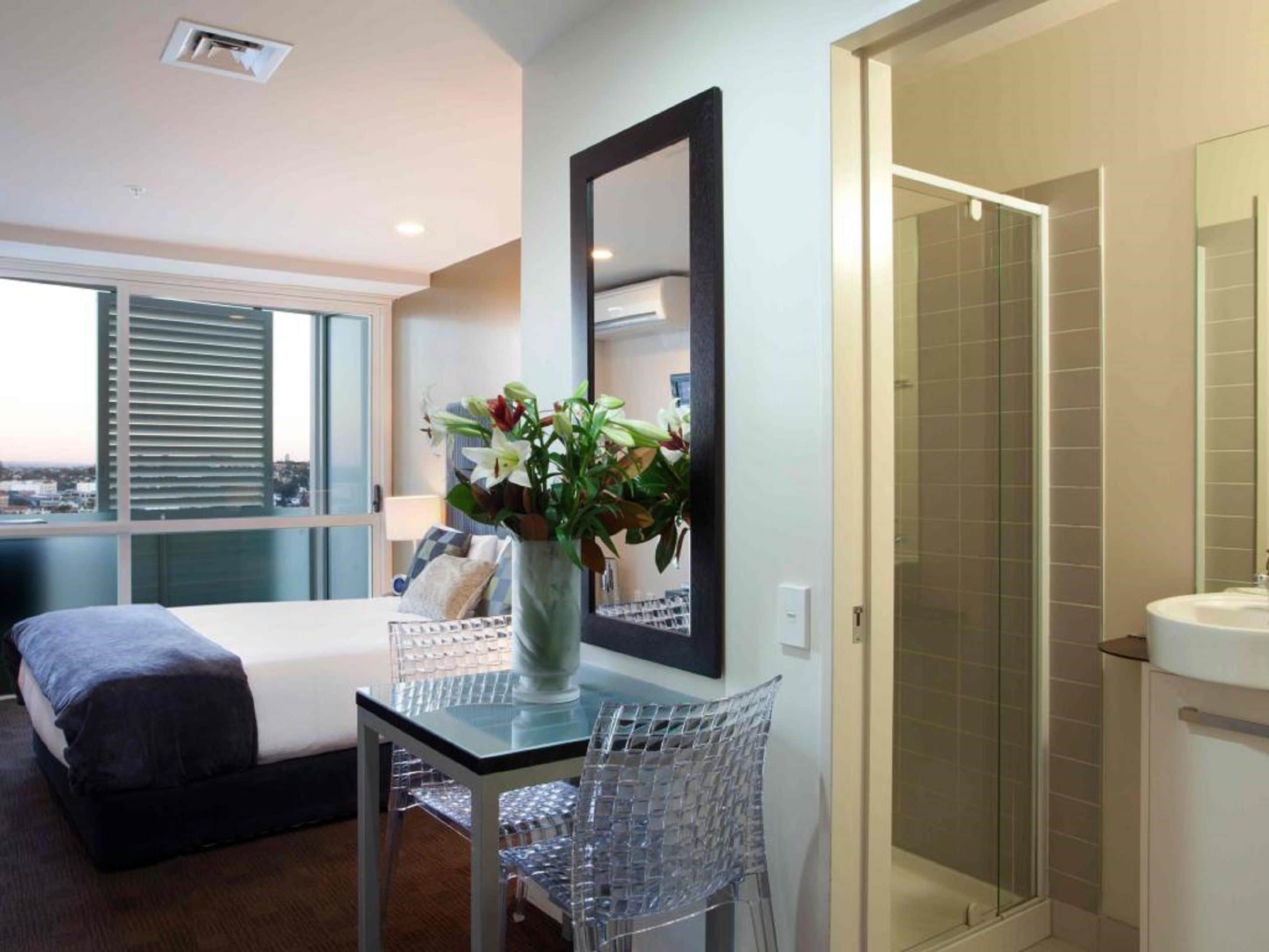 Ett modernt hotellrum med en säng, ett glasbord med en vas med blommor, en stor spegel och en duschhörna.