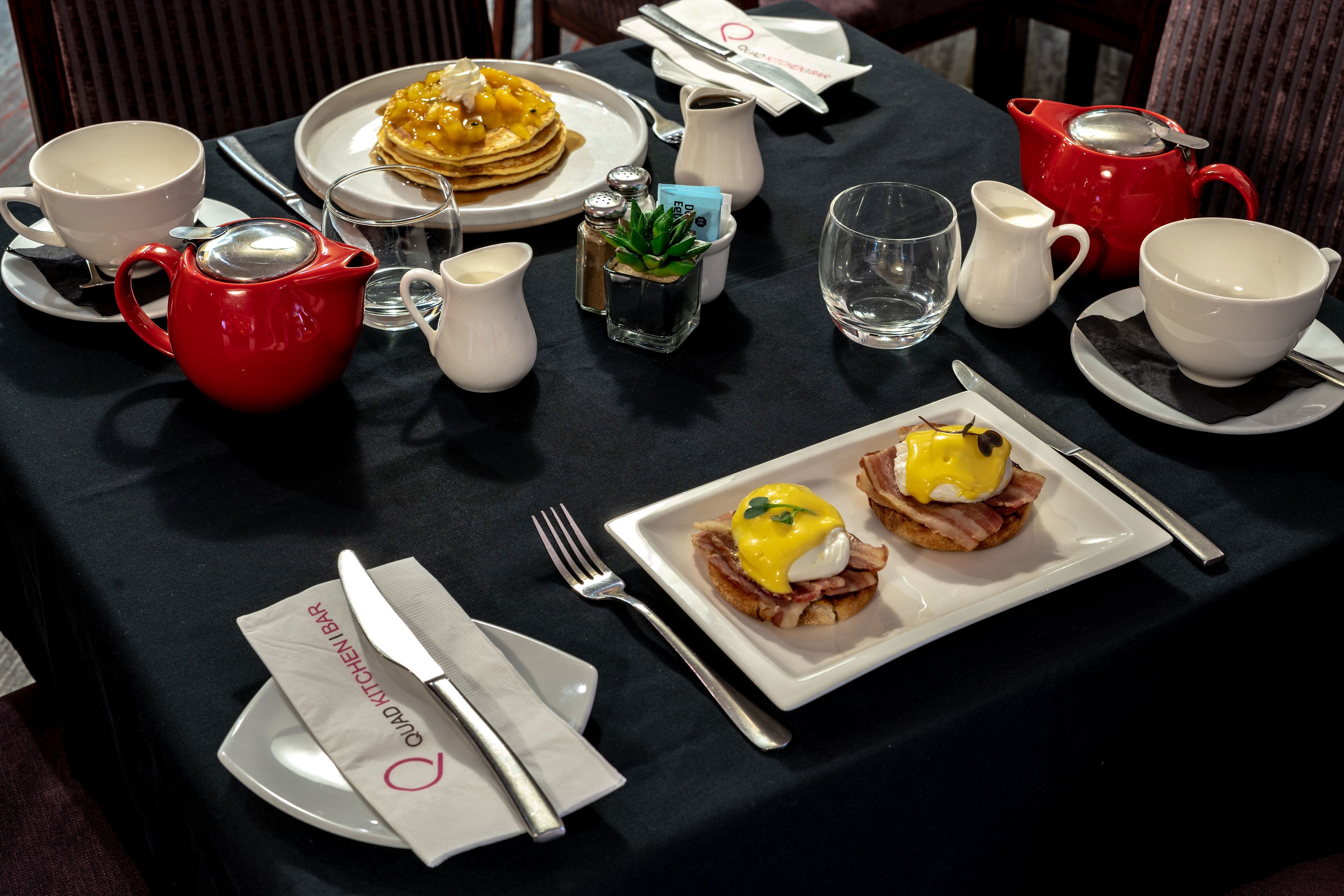 A breakfast table with plates of food, cups, a red teapot, and a small plant.