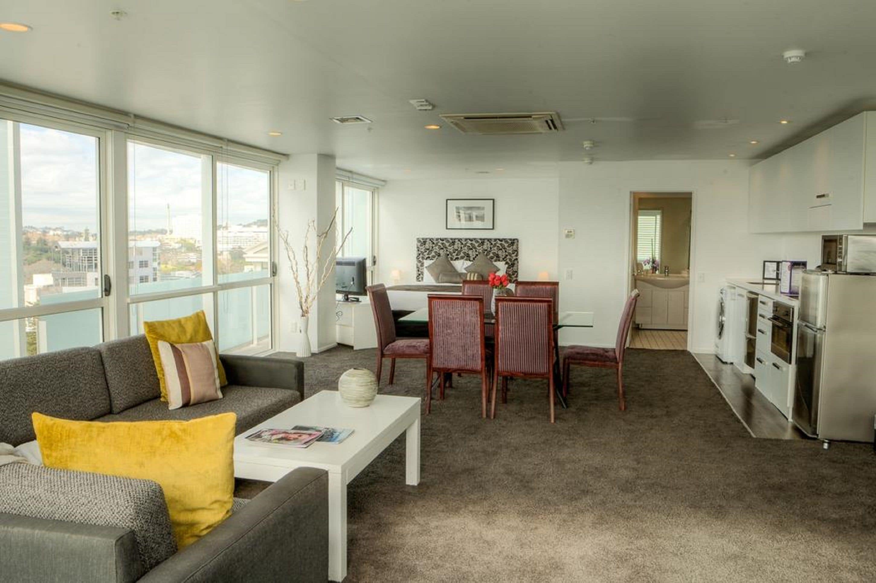 Una sala de estar moderna con un sofá gris, una mesa de centro y un comedor con mesa y sillas. Hay una cocina con un refrigerador y un microondas.