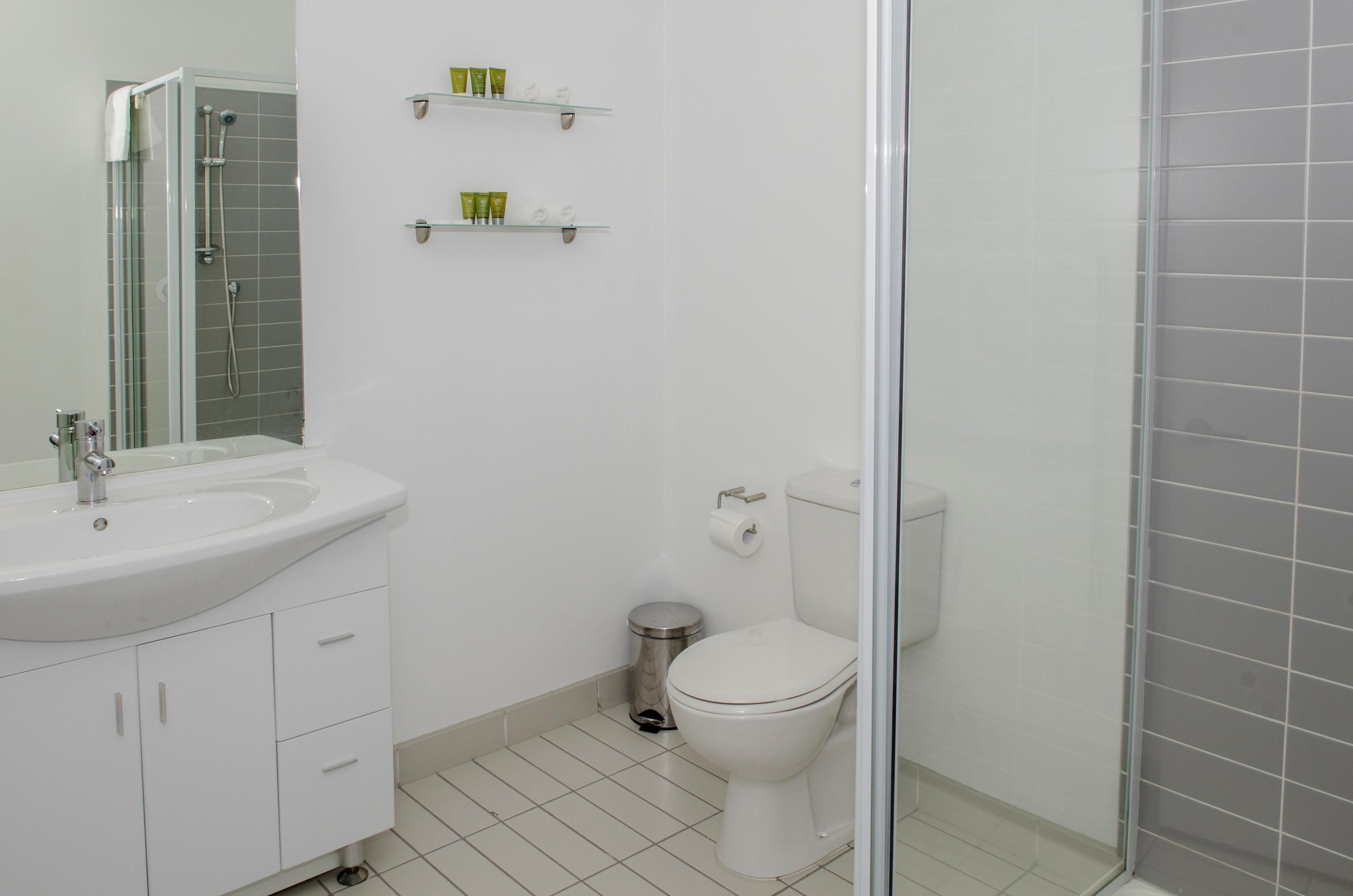 A bathroom with a white sink, mirror, toilet, and shower. There are shelves with toiletries.