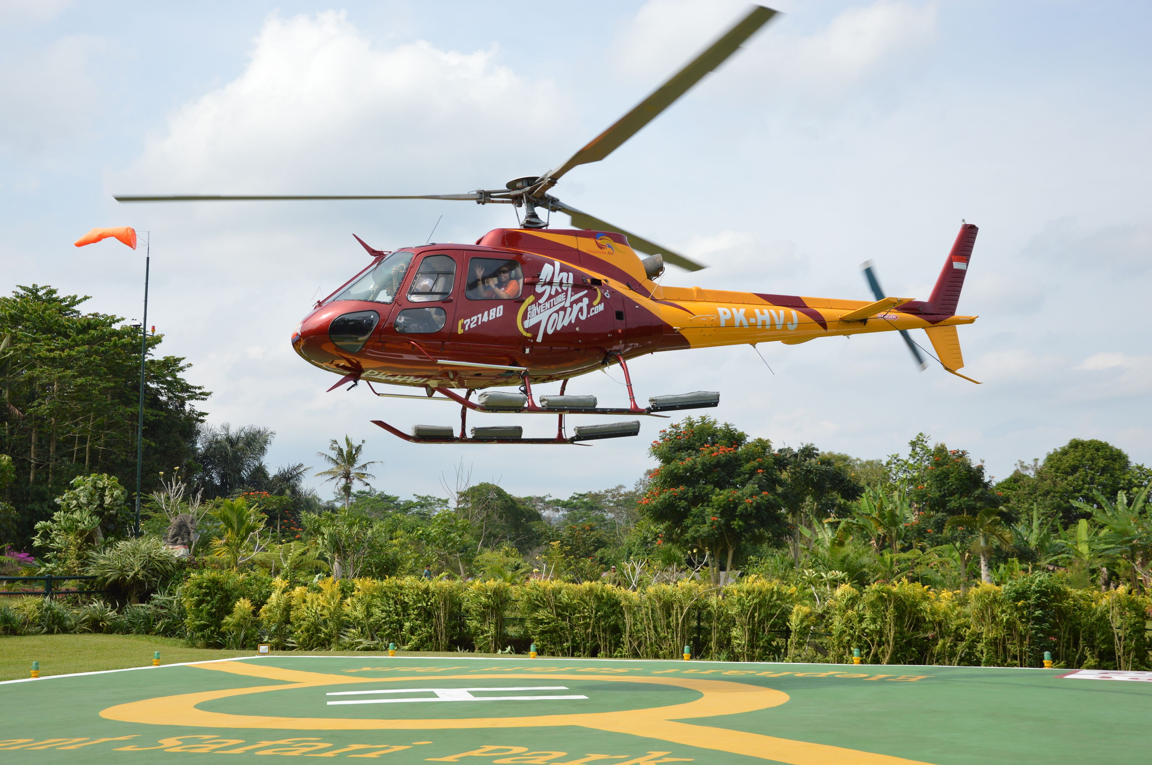 A red and yellow helicopter with "Sky Tours" branding, hovering over a green field with a large yellow and green logo.