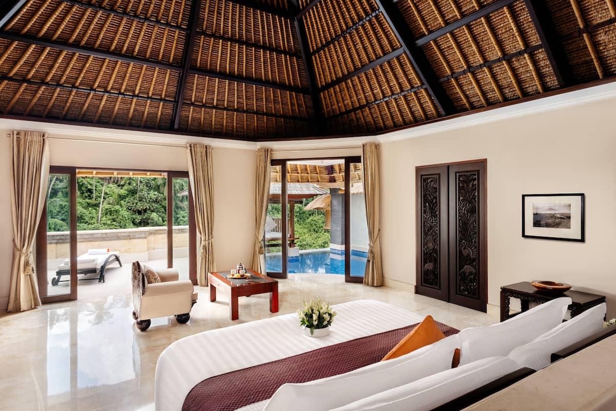 A spacious bedroom with a large bed, a sitting area with a table and chairs, and a view of a swimming pool and lush greenery.