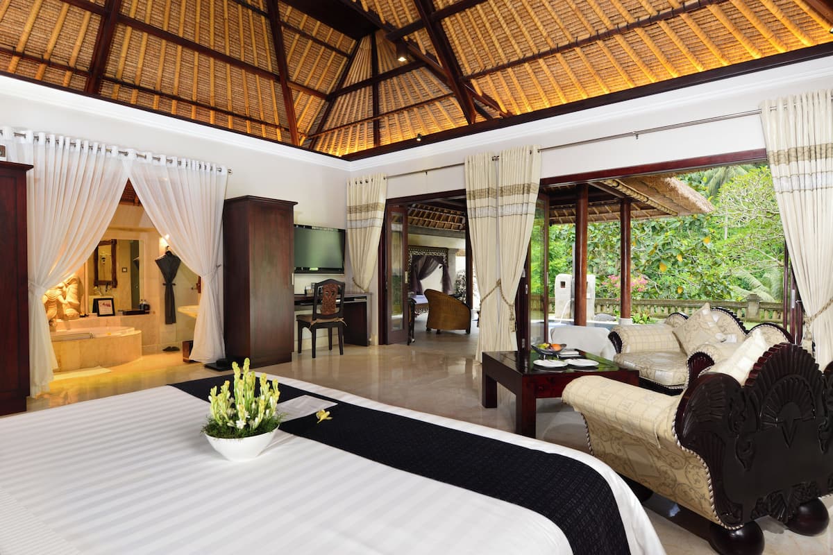A spacious room with a large bed, a sofa, a television, and a desk. There is a view of greenery outside the windows.