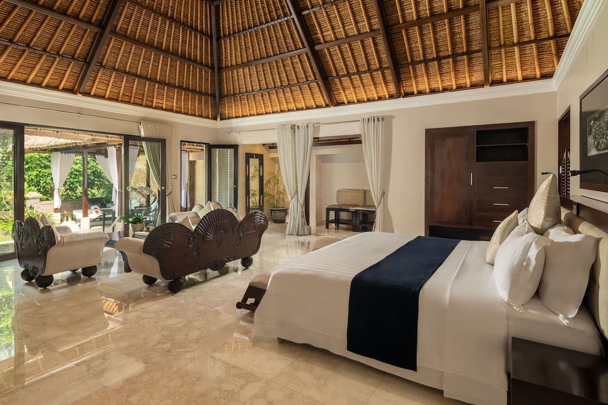A spacious bedroom with a large bed, a seating area with sofas, and a view of the outdoors through large windows.