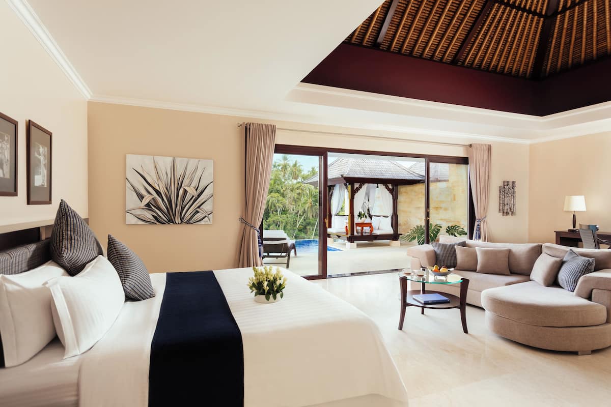 A hotel room with a large bed, a sofa, a glass coffee table, and a view of a pool and outdoor area.