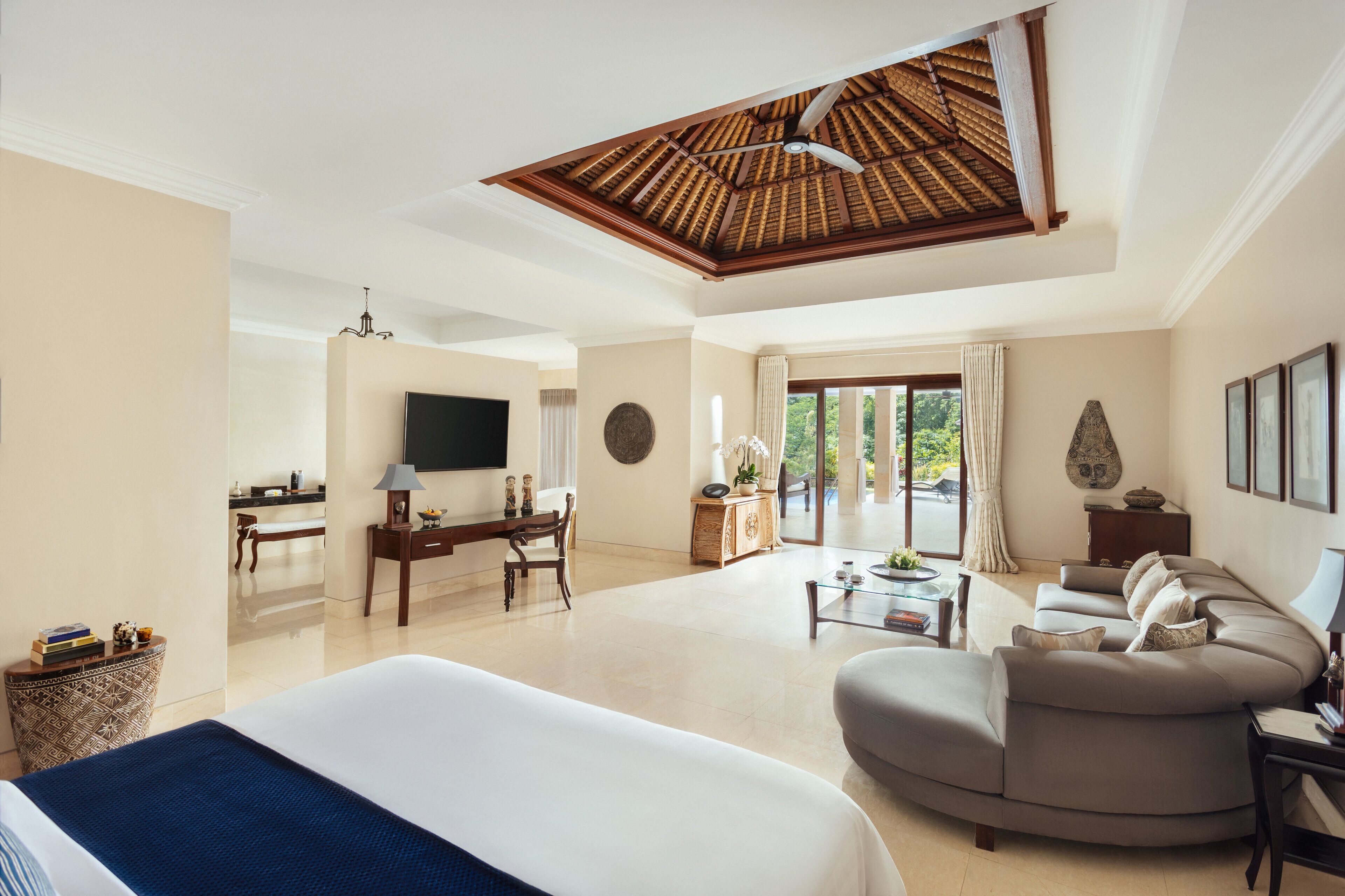 A spacious hotel room with a large bed, a curved sofa, a dining area, and a ceiling fan.