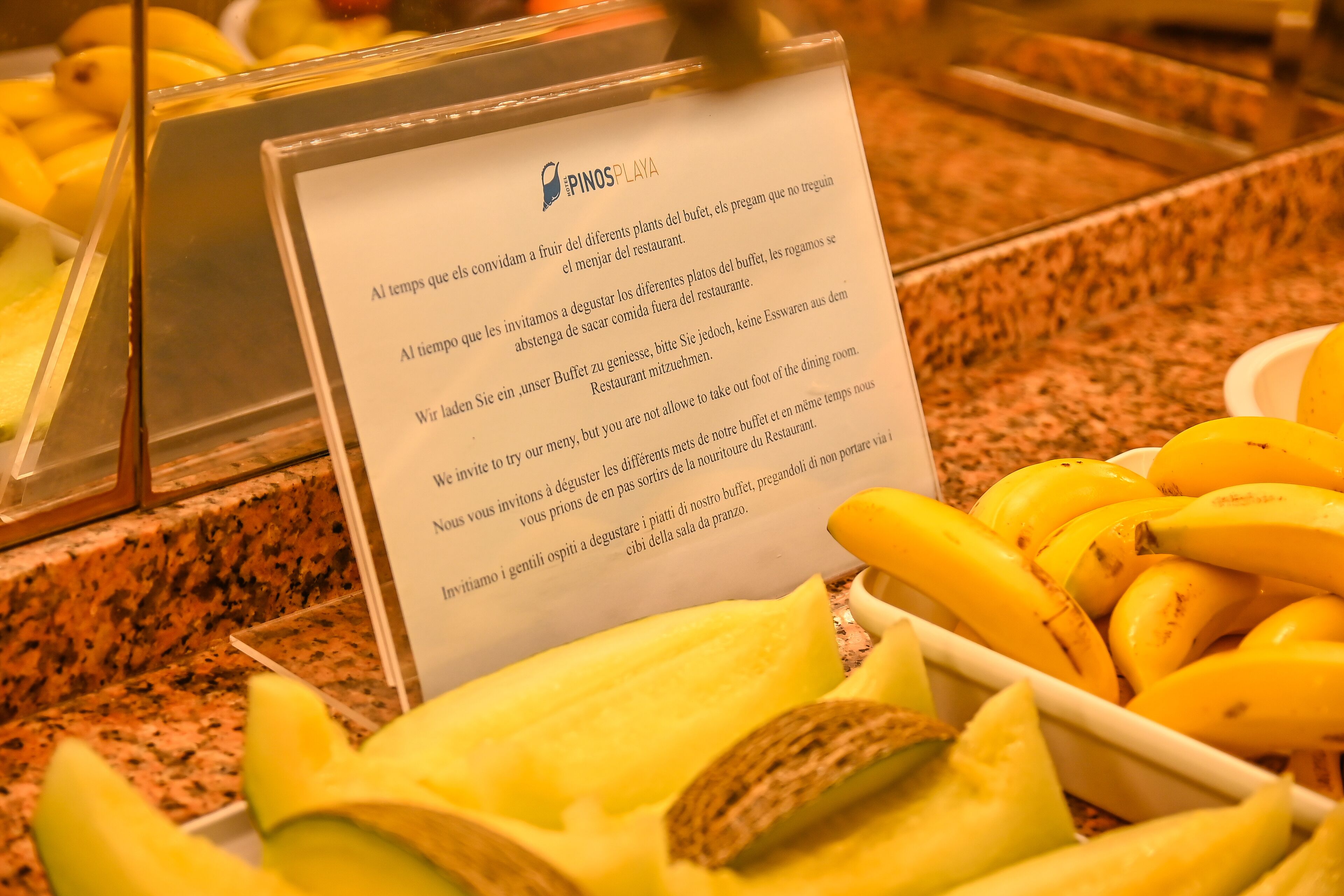 A buffet area with a sign in Spanish, bananas, and a container of sliced fruit.