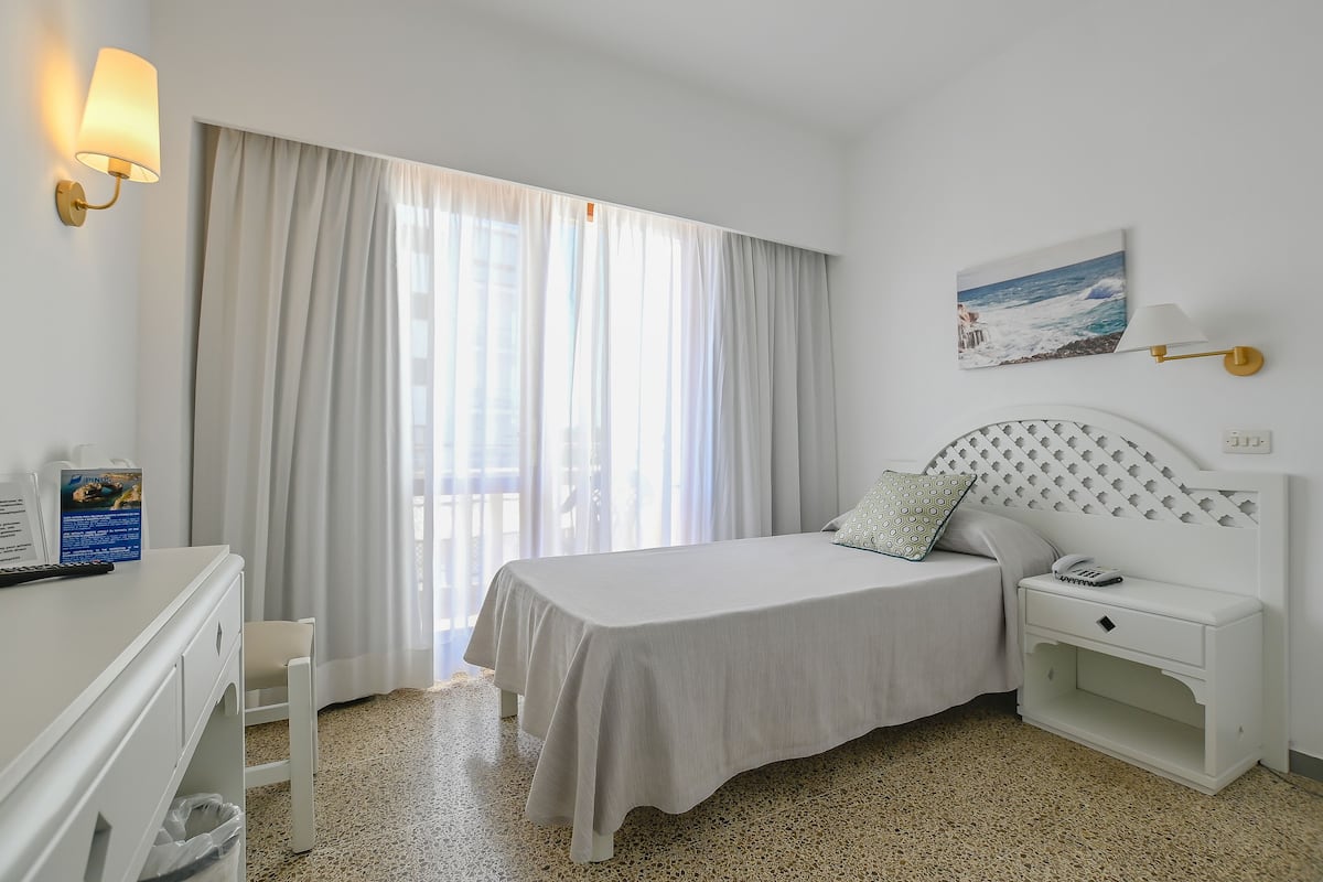 A hotel room with a bed, bedside table, lamp, and a painting of a beach scene.